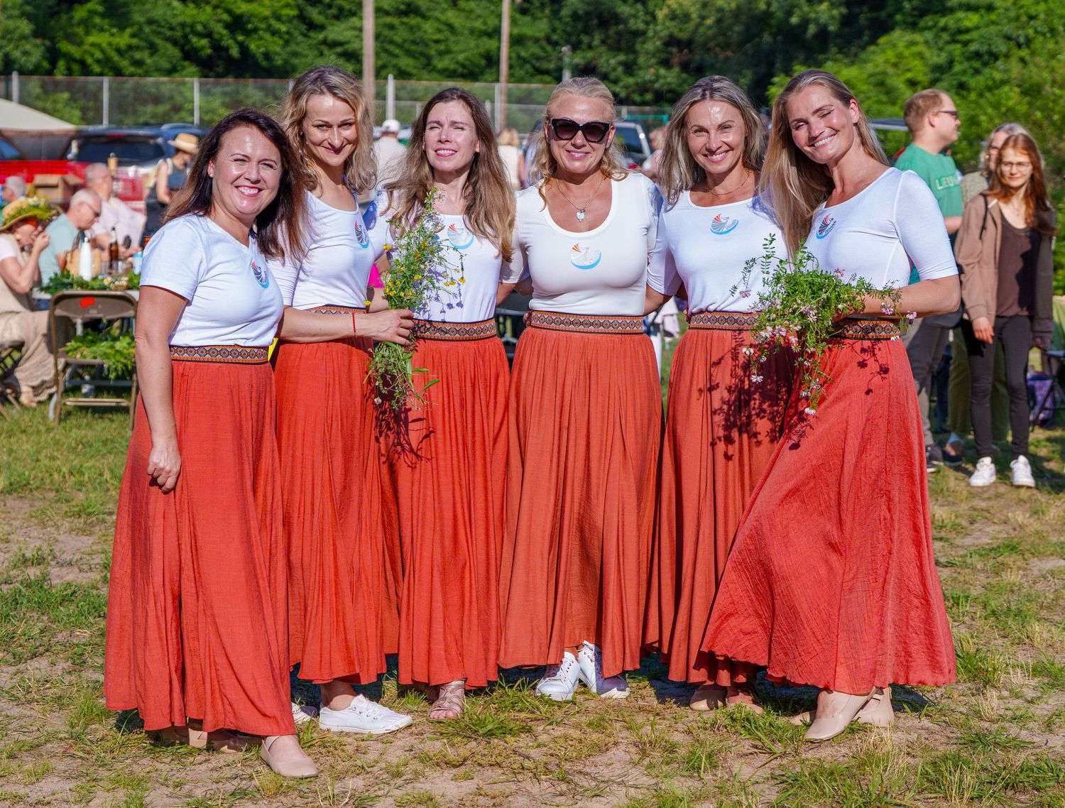Six women in matching red skirts and white tops pose together outdoors holding wildflowers at a summer festival.