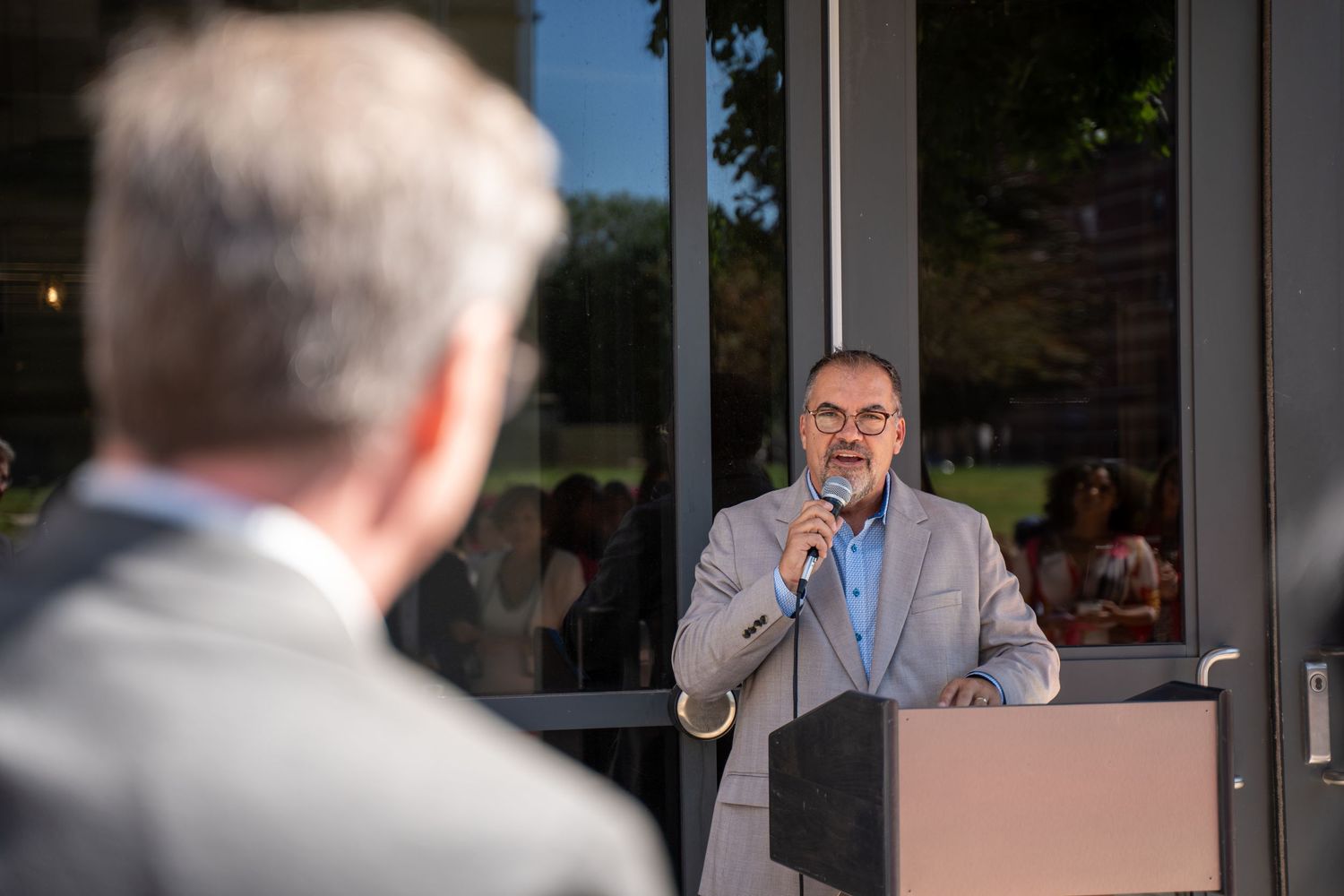 Man in gray suit speaking at podium with microphone at formal outdoor evening event.