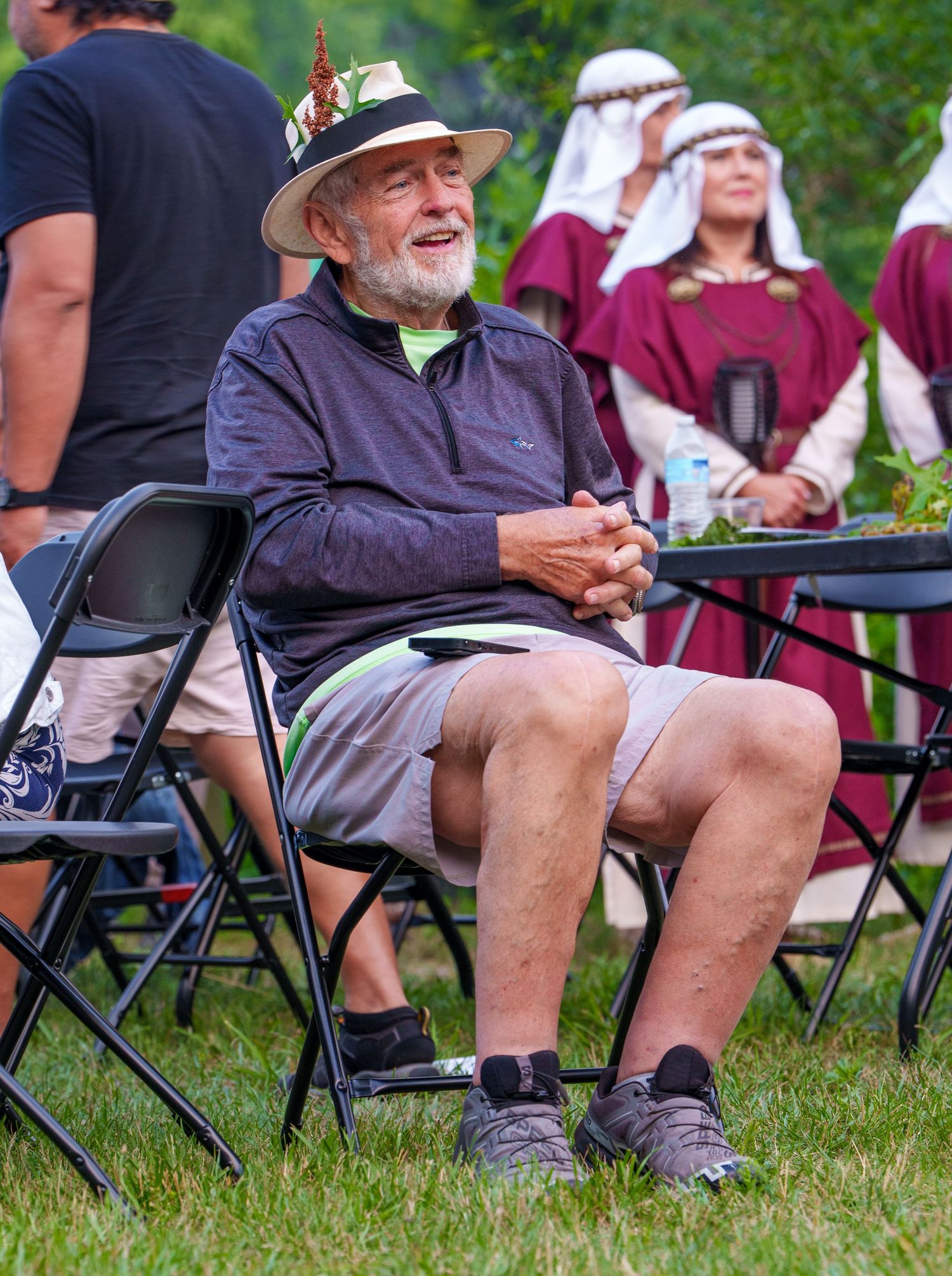 Smiling elderly man in a hat and shorts sitting in a folding chair at an outdoor medieval festival event.