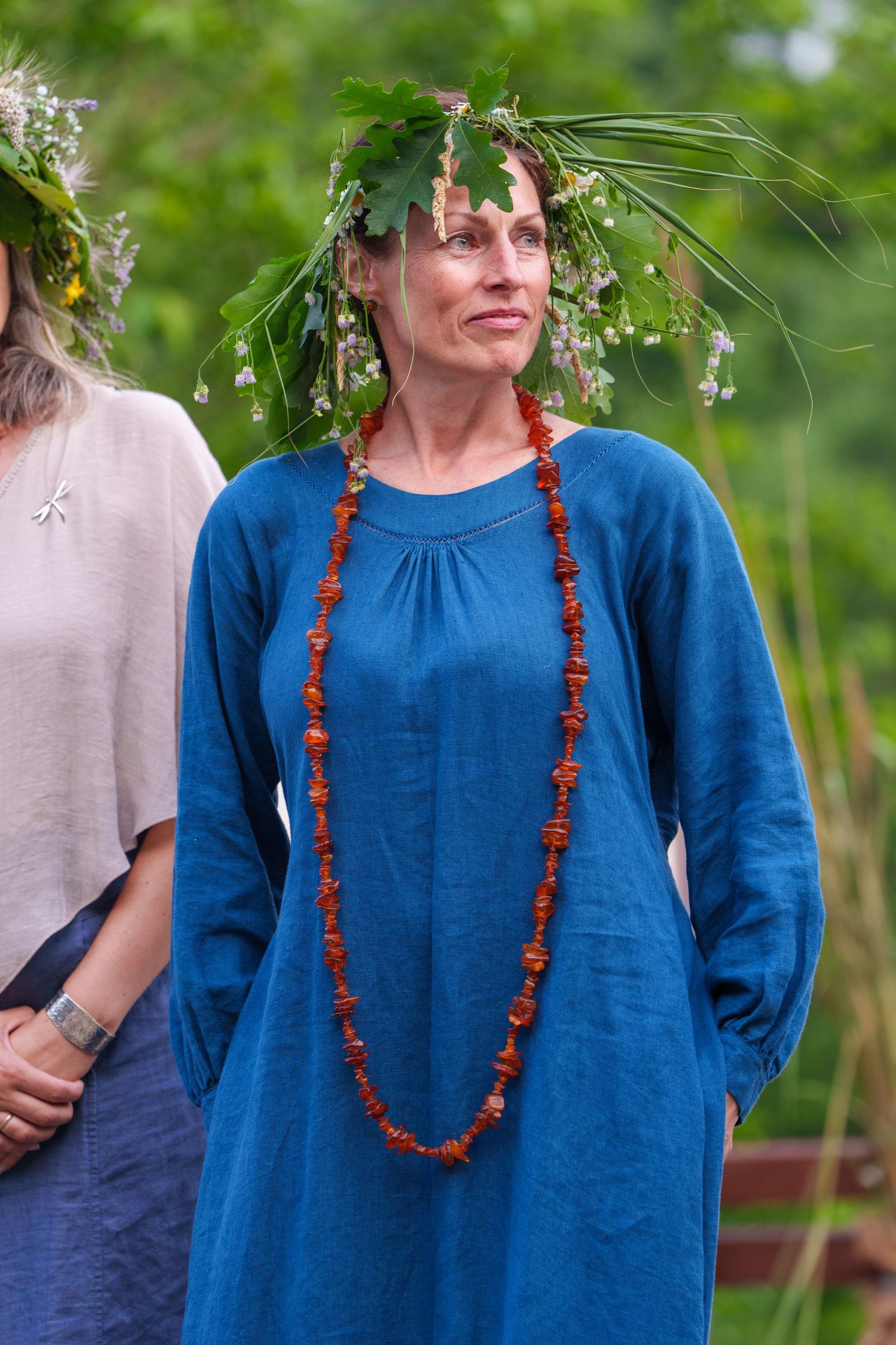 Woman in blue linen dress with amber necklace and floral crown at outdoor summer festival.