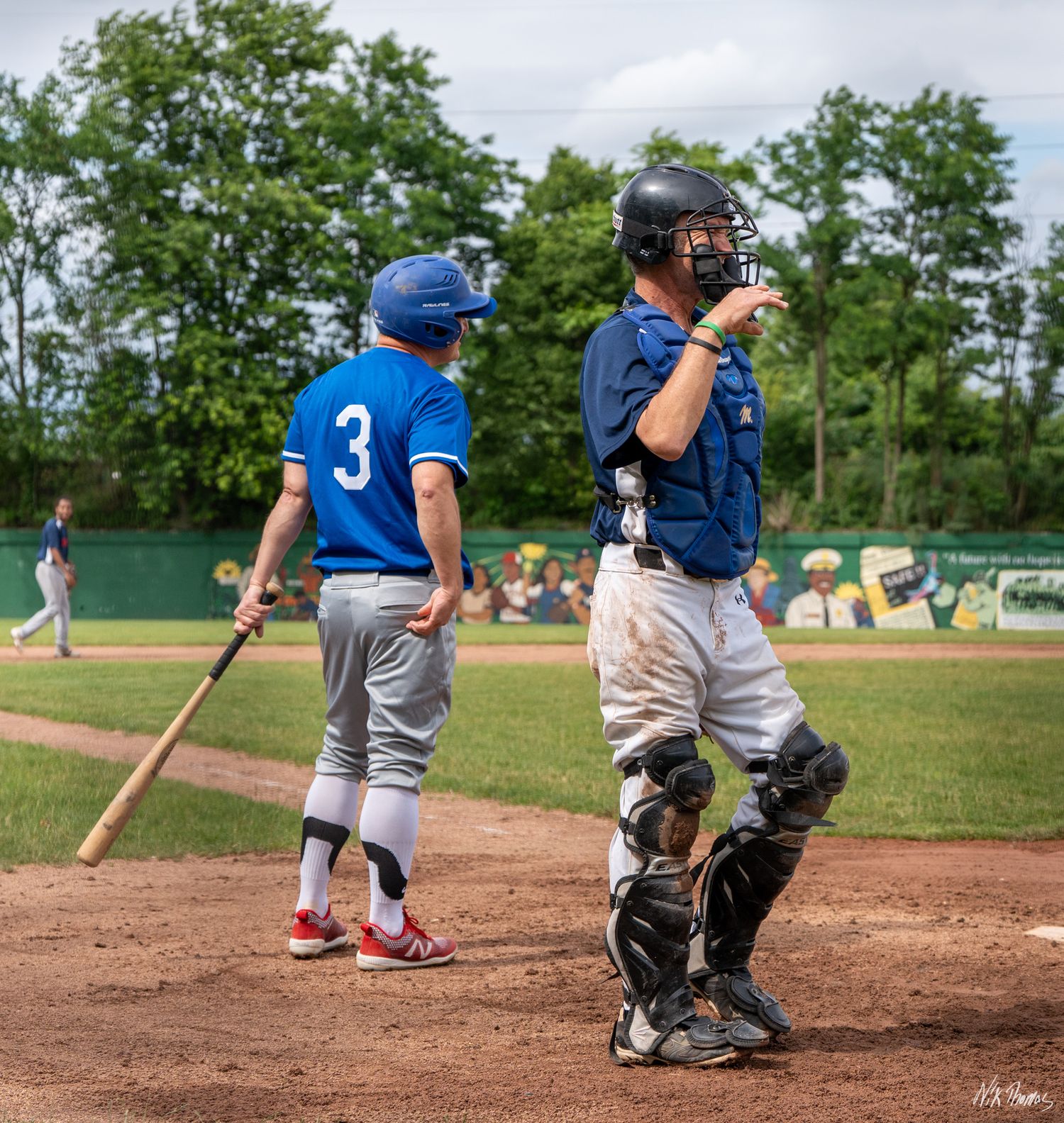 Baseball catcher in gear stands near batter wearing number 3 blue jersey during a youth baseball game outdoors.