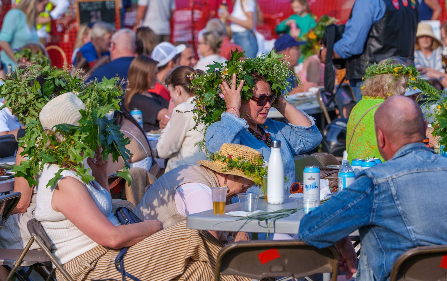 People wearing floral wreaths celebrate Midsummer festival outdoors at sunny tables.