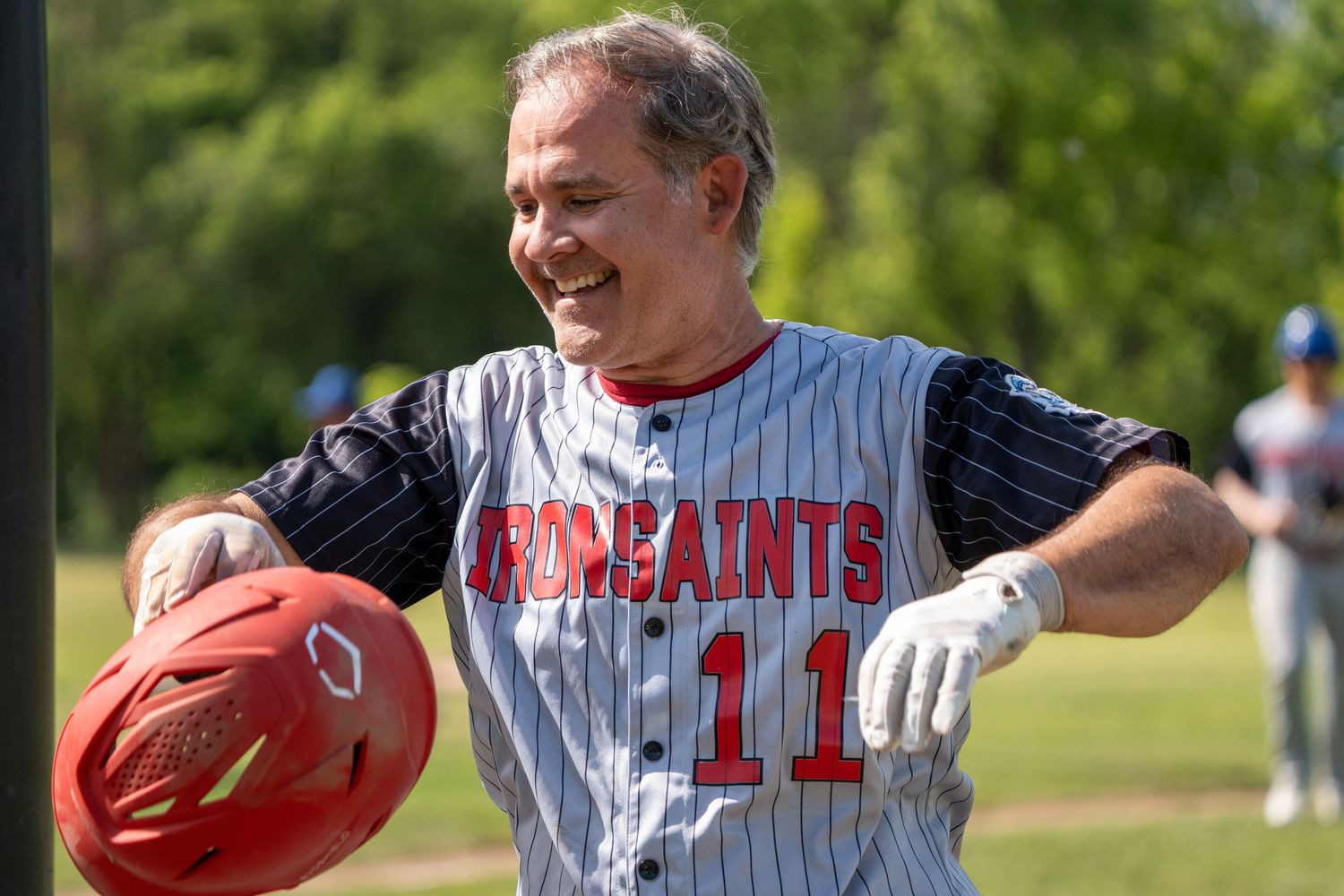 Smiling baseball player wearing Saints #11 jersey holds red helmet on sunny outdoor field.