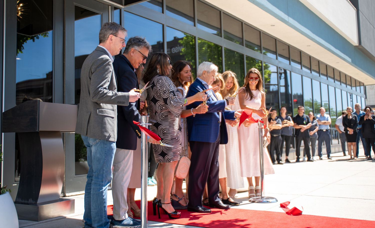 Group of people cutting a red ribbon at a modern building grand opening ceremony outdoors.