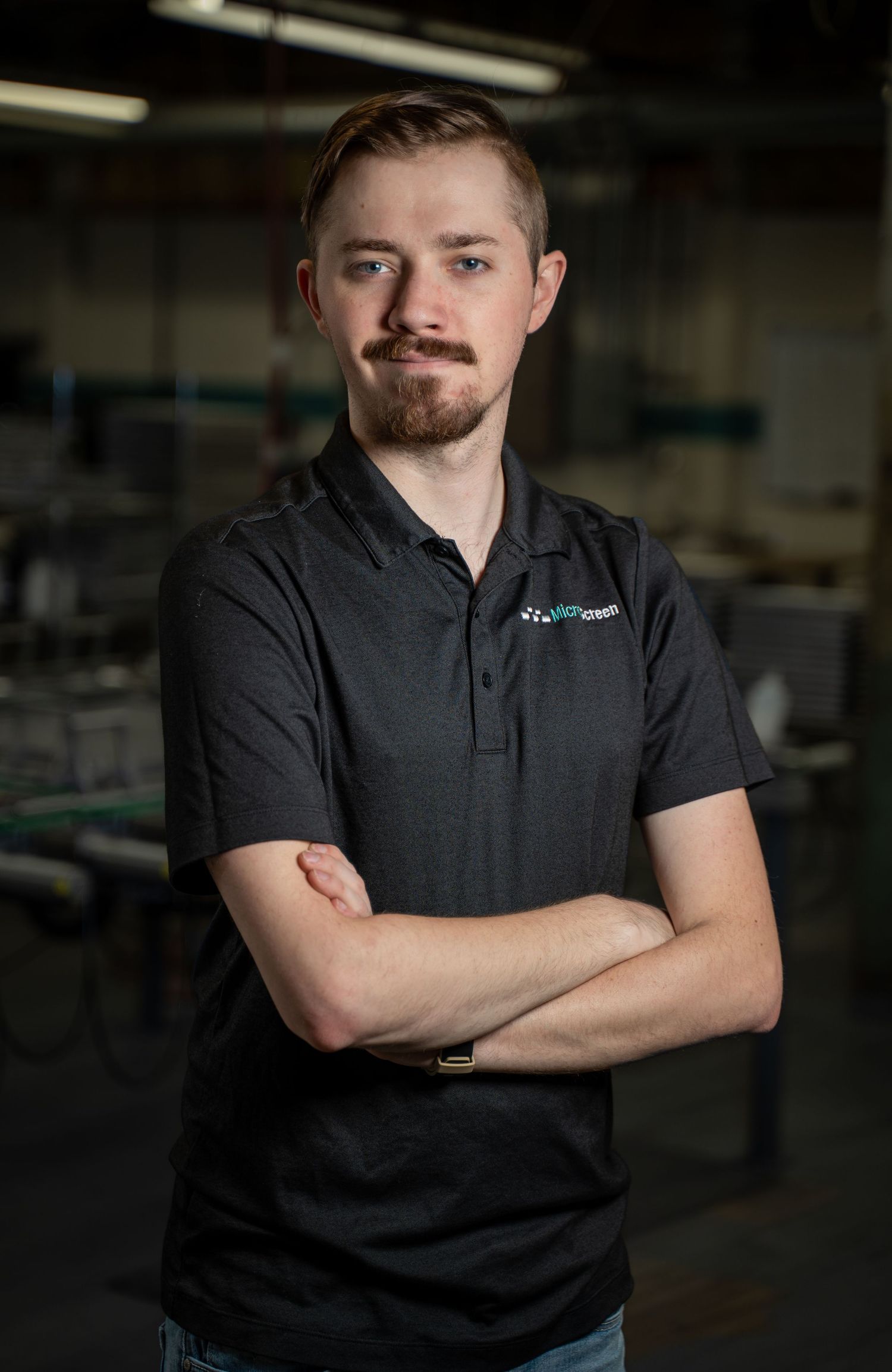 Young man with beard in black polo shirt stands with arms crossed in an industrial workspace.