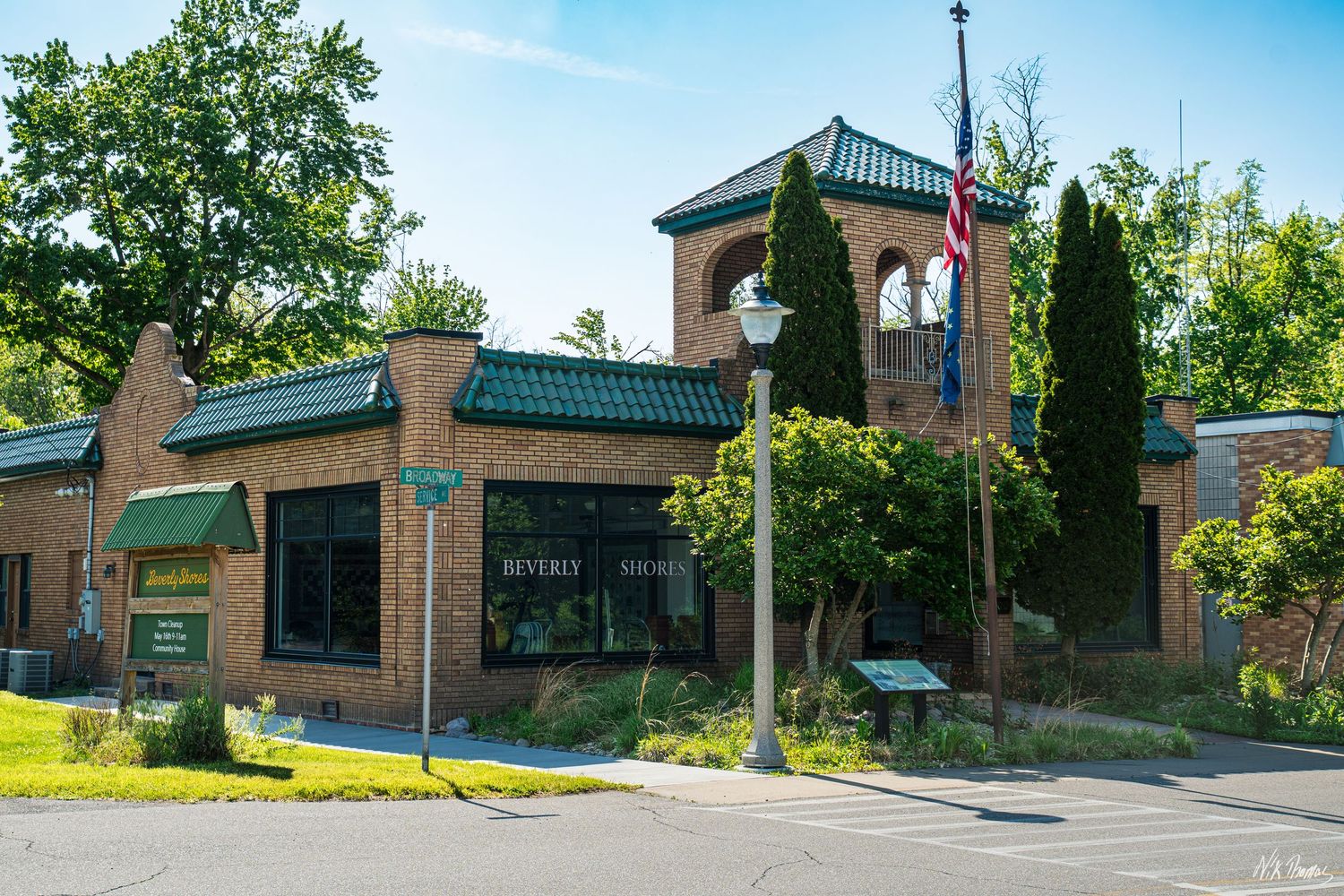 Brick building with green roof accents, American flag, tall cypress trees, and manicured landscaping on a sunny day.