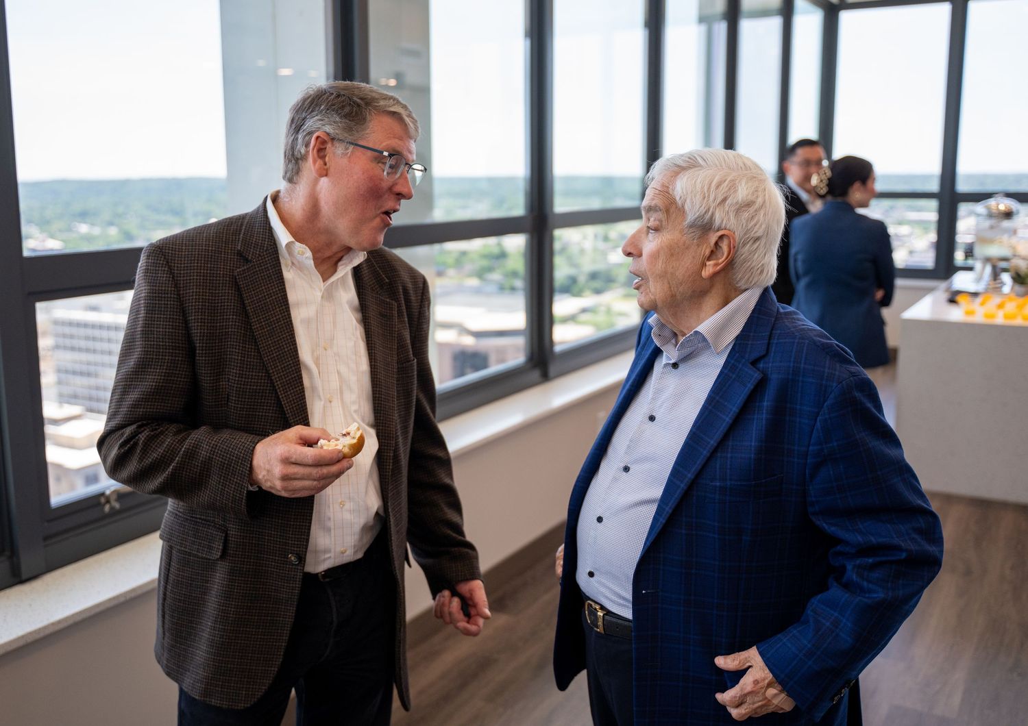 Two businessmen in blazers engage in conversation near large windows with a city skyline view.