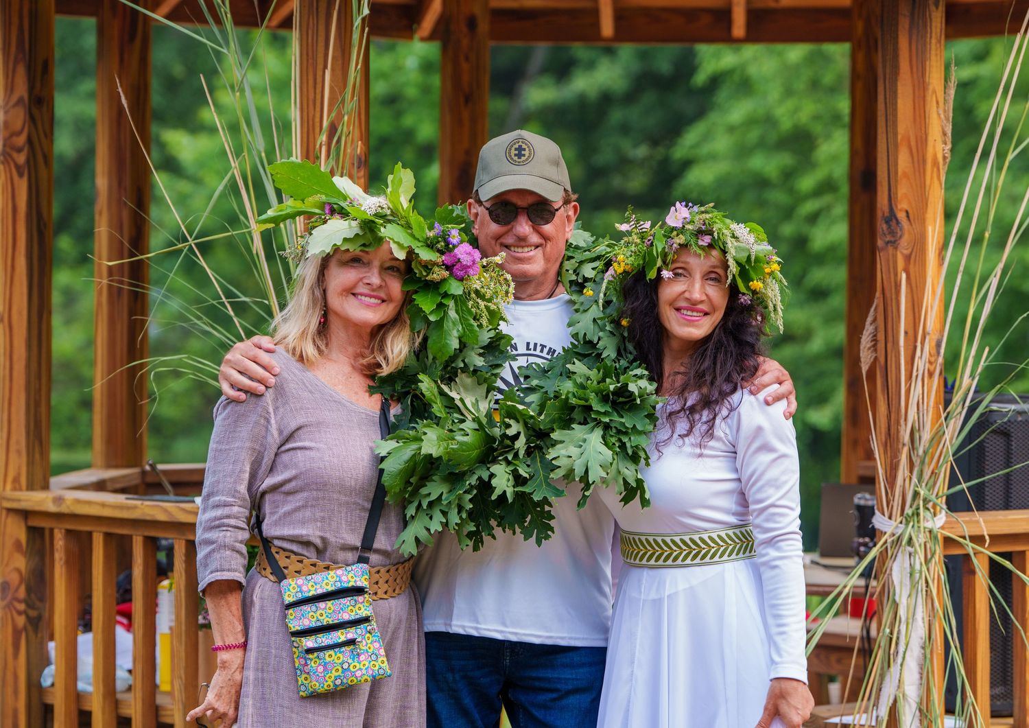 Three people wearing floral wreaths smile together on a wooden deck surrounded by lush greenery.