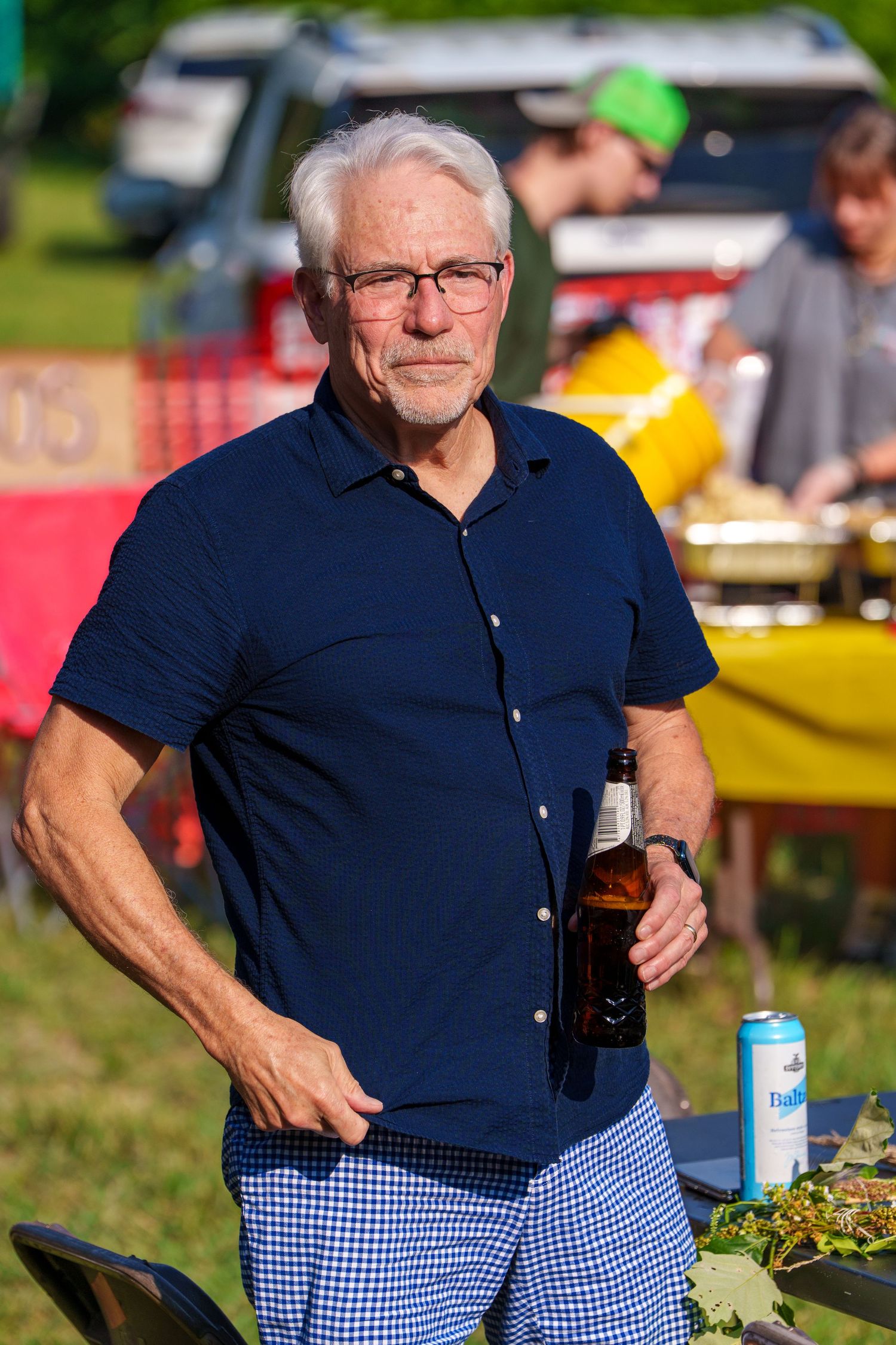 Senior man with white hair and glasses holding beer bottle at outdoor summer gathering.