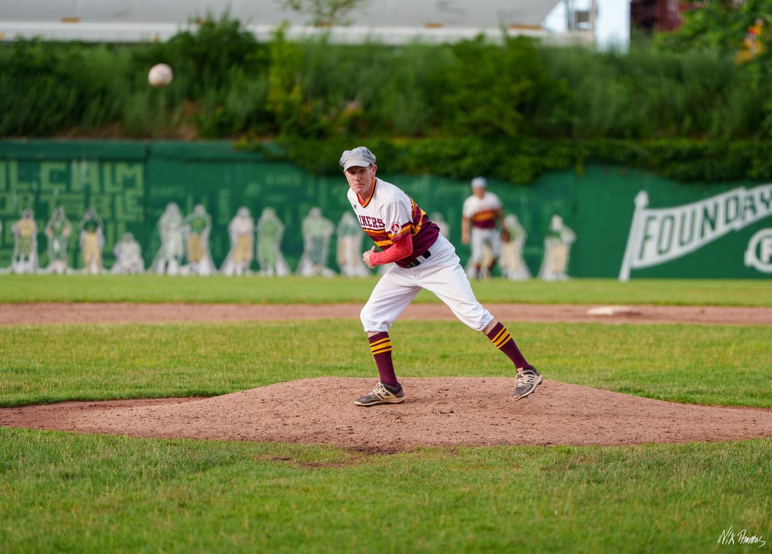 Baseball pitcher in white and maroon uniform mid-windup on mound during game with green outfield wall behind.