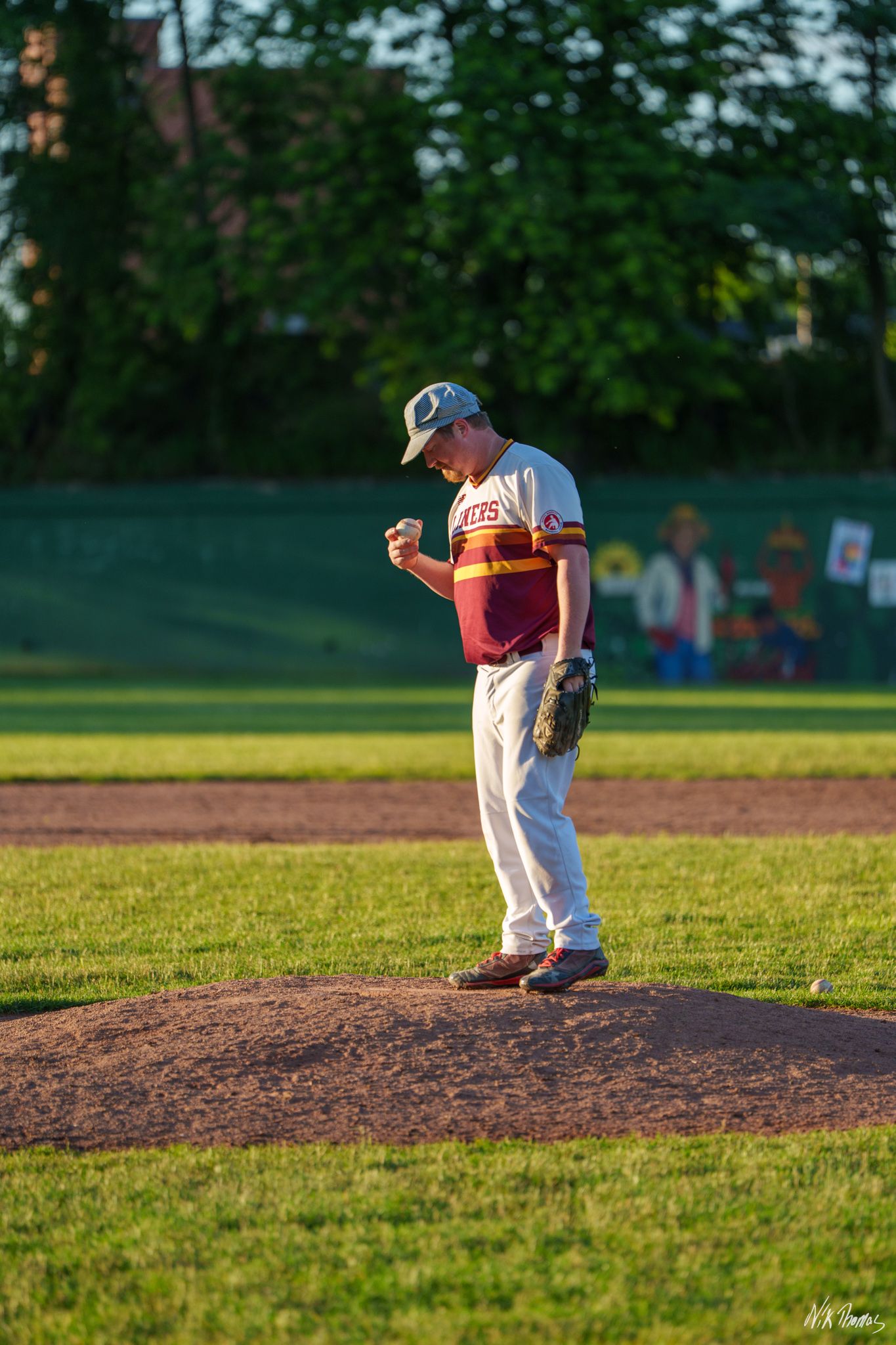 A baseball pitcher in maroon and white uniform stands on the mound preparing to pitch during a game.