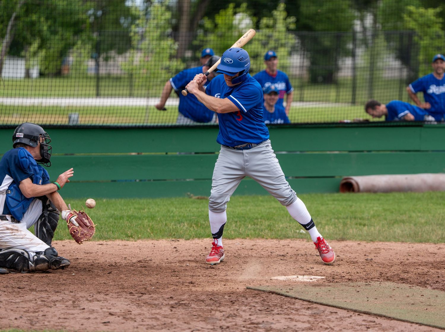 Baseball batter in blue uniform swings at pitch while catcher crouches behind home plate during outdoor game.
