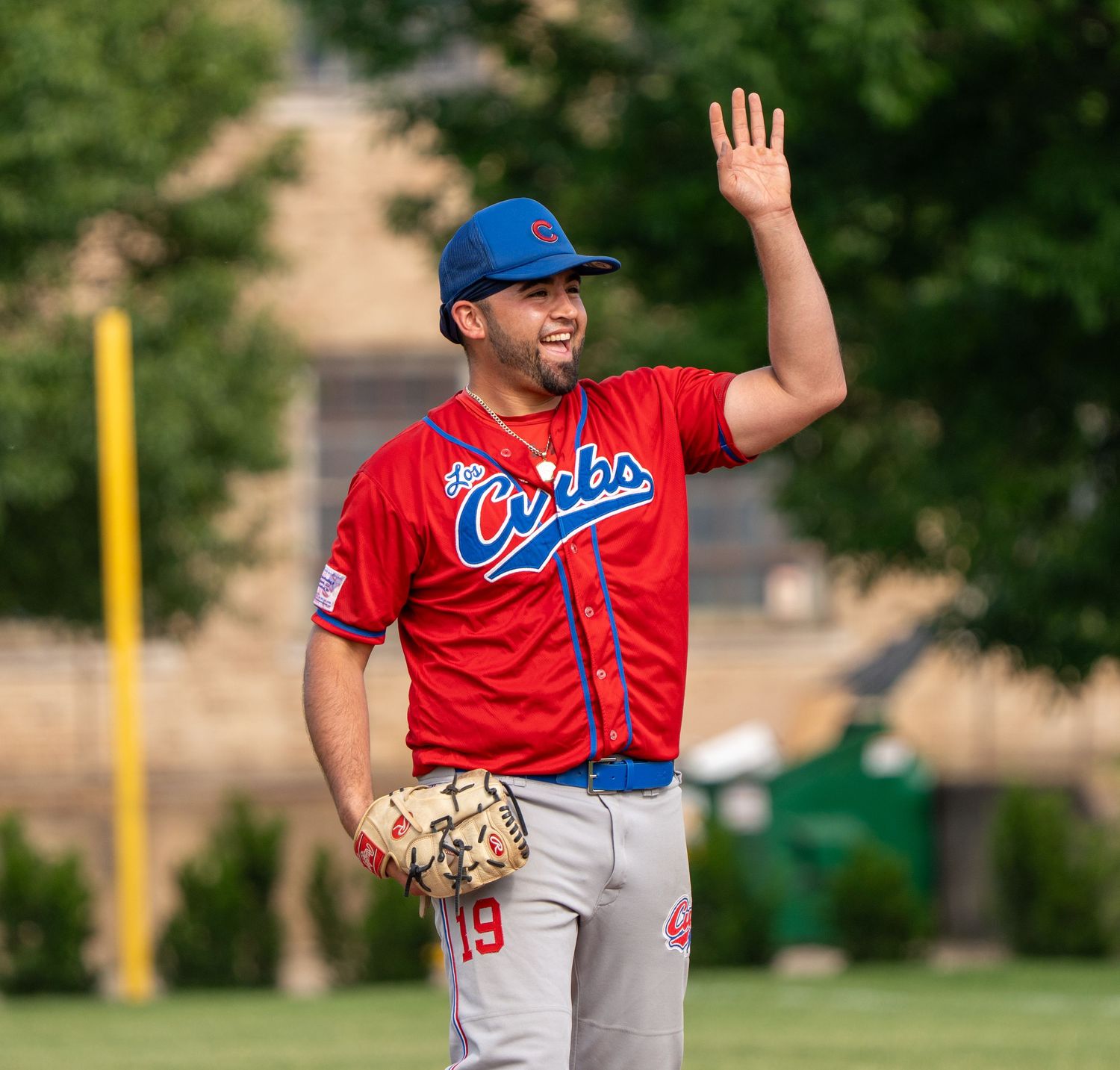 Baseball player in red Cubs jersey number 19 waves while holding glove on sunny green field.