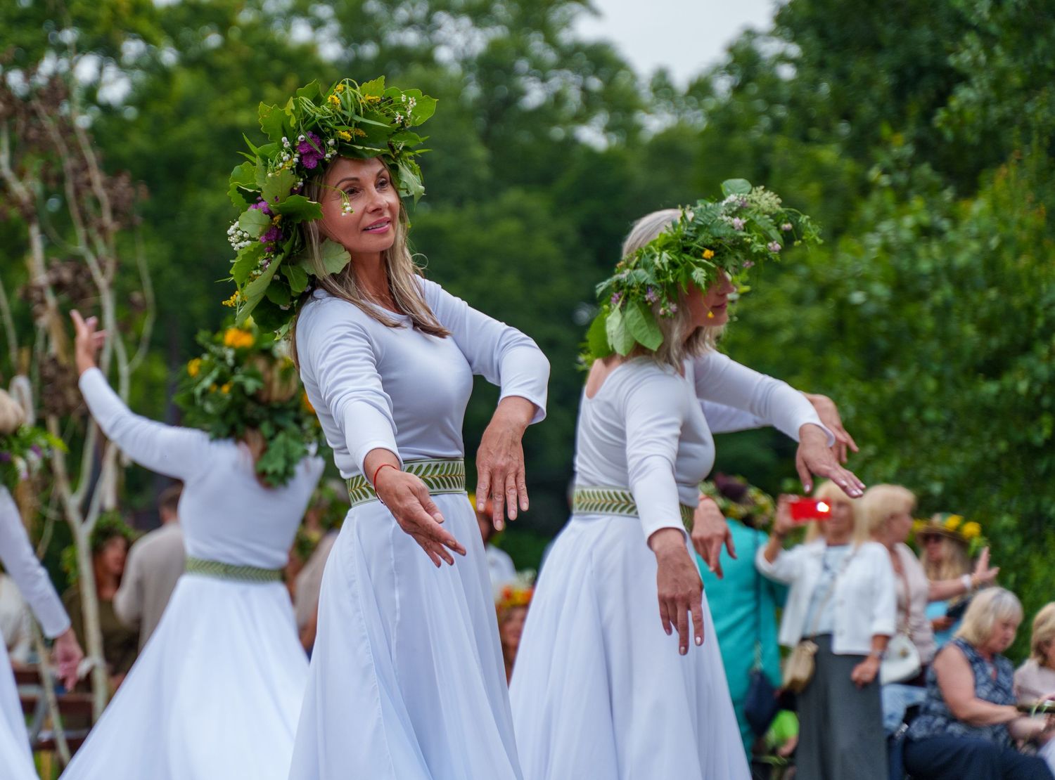 Women in white dresses and floral wreaths perform a traditional folk dance outdoors at a summer festival.