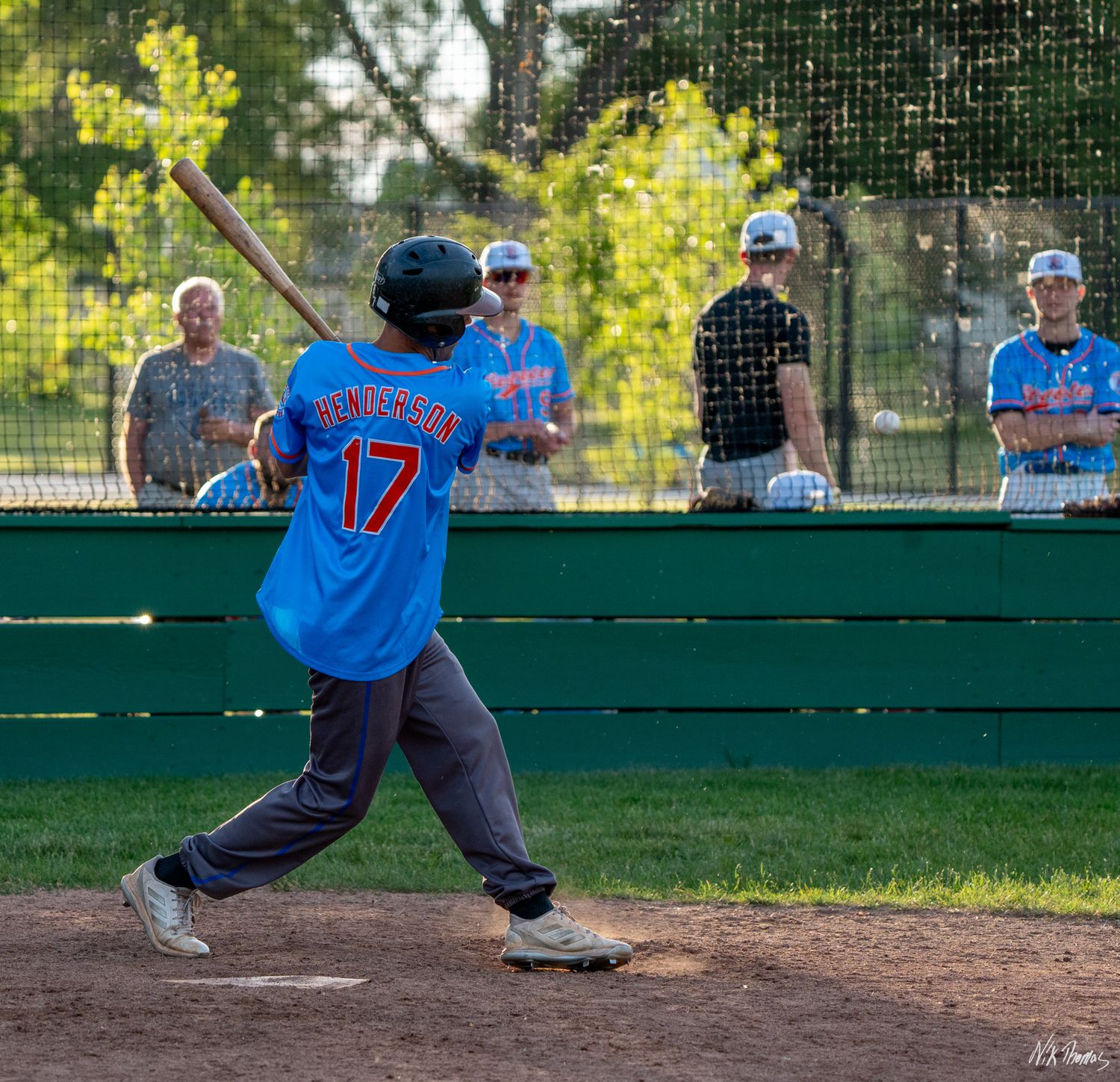 Baseball batter in blue jersey number 17 swings at pitch during game with teammates watching from dugout.