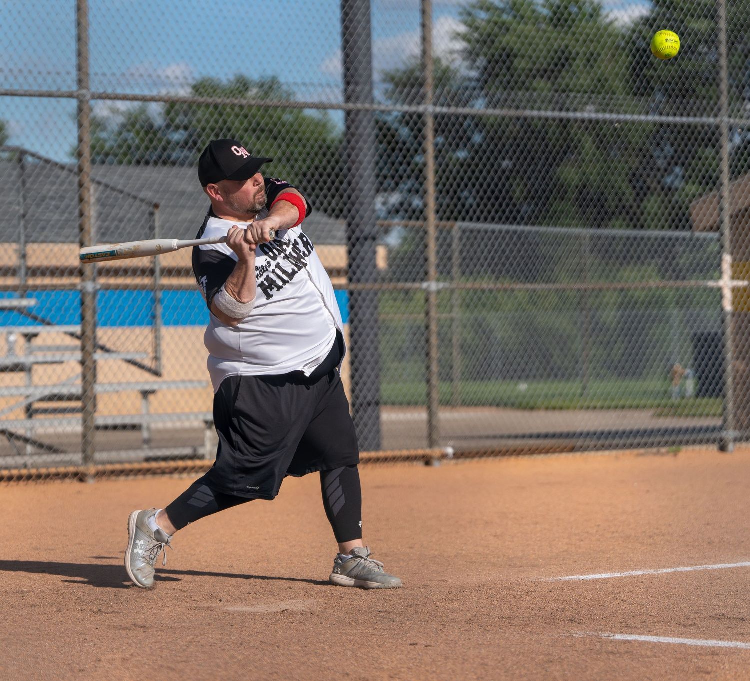 A batter swings at a yellow softball during an outdoor game on a sunny day at a chain-link fenced diamond.