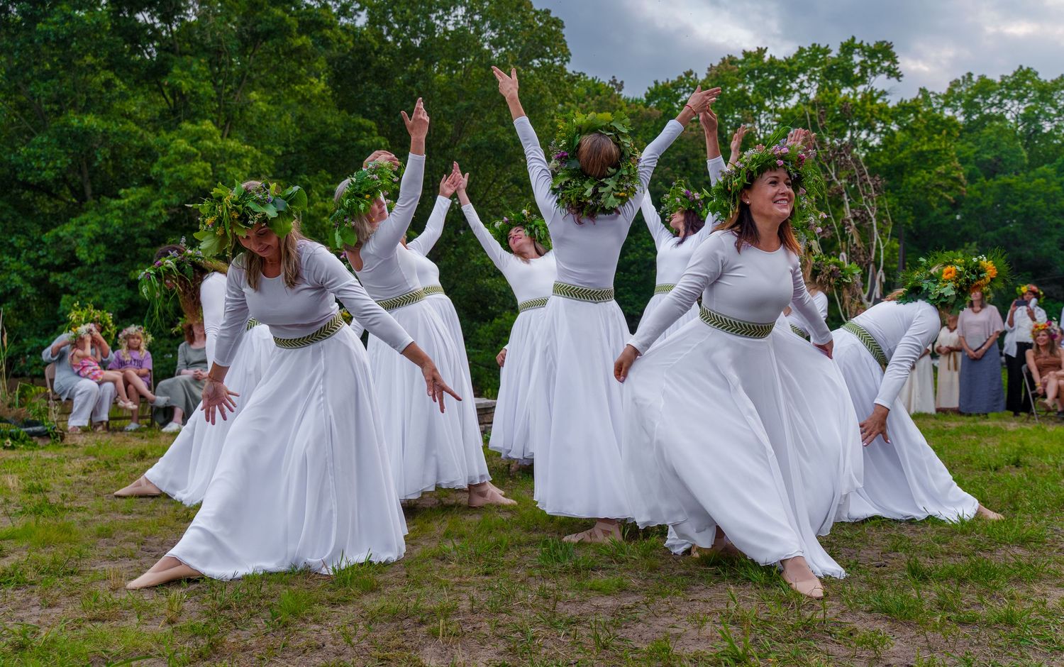 Women in white dresses and floral crowns perform an elegant outdoor dance ceremony in a lush green forest setting.