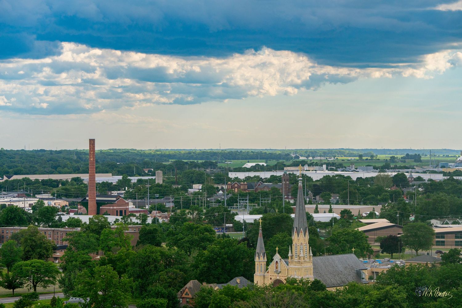Aerial view of a small city skyline with a Gothic church, brick smokestack, and lush green trees under dramatic storm clouds.
