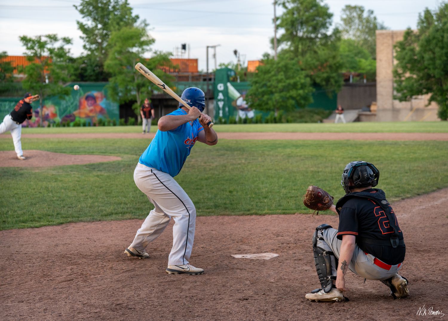 Baseball batter in blue jersey prepares to swing at pitch while catcher crouches behind home plate on sunny day.