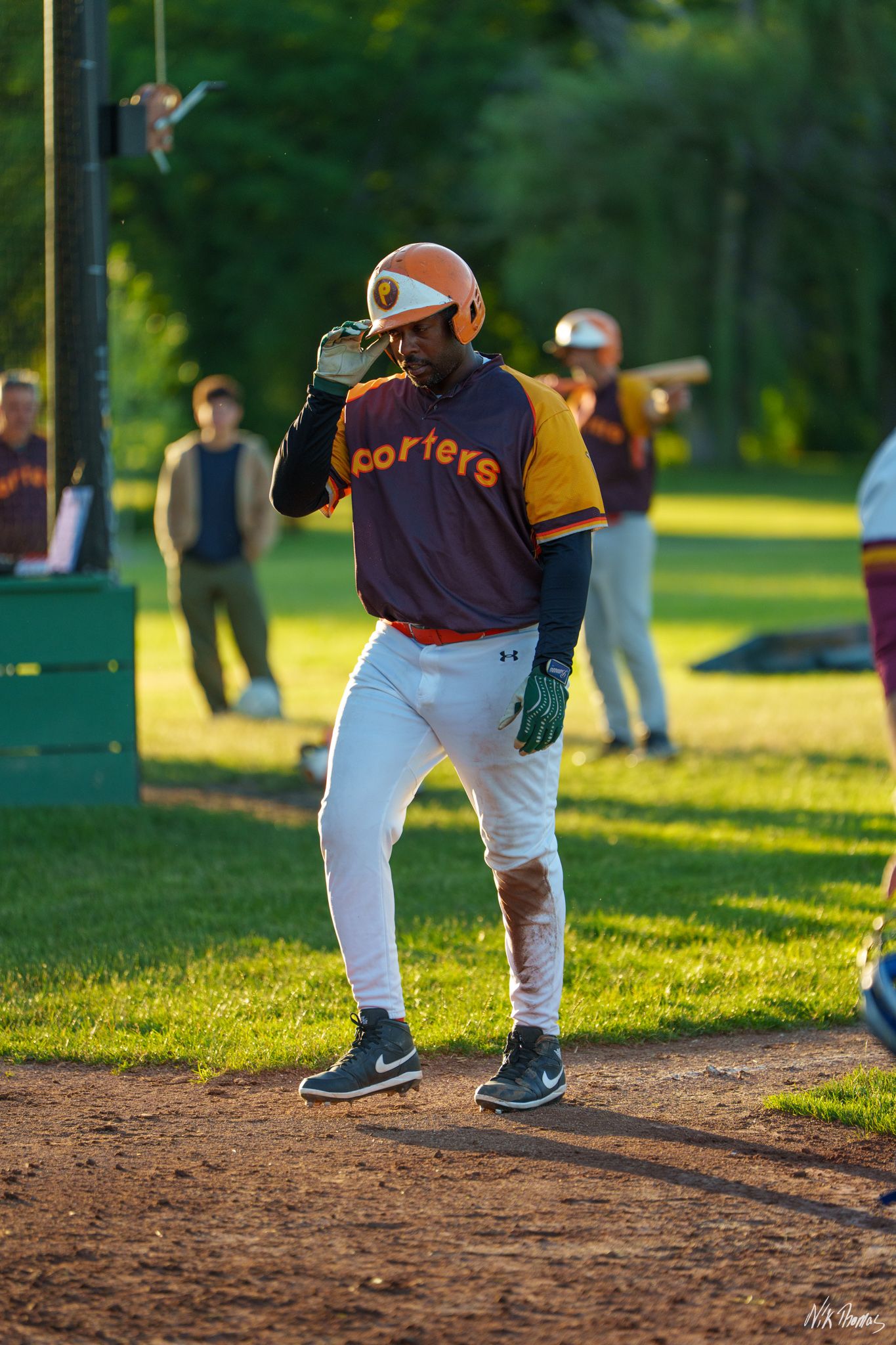 Baseball player in Corters uniform adjusting orange helmet on dirt base path during sunny outdoor game.