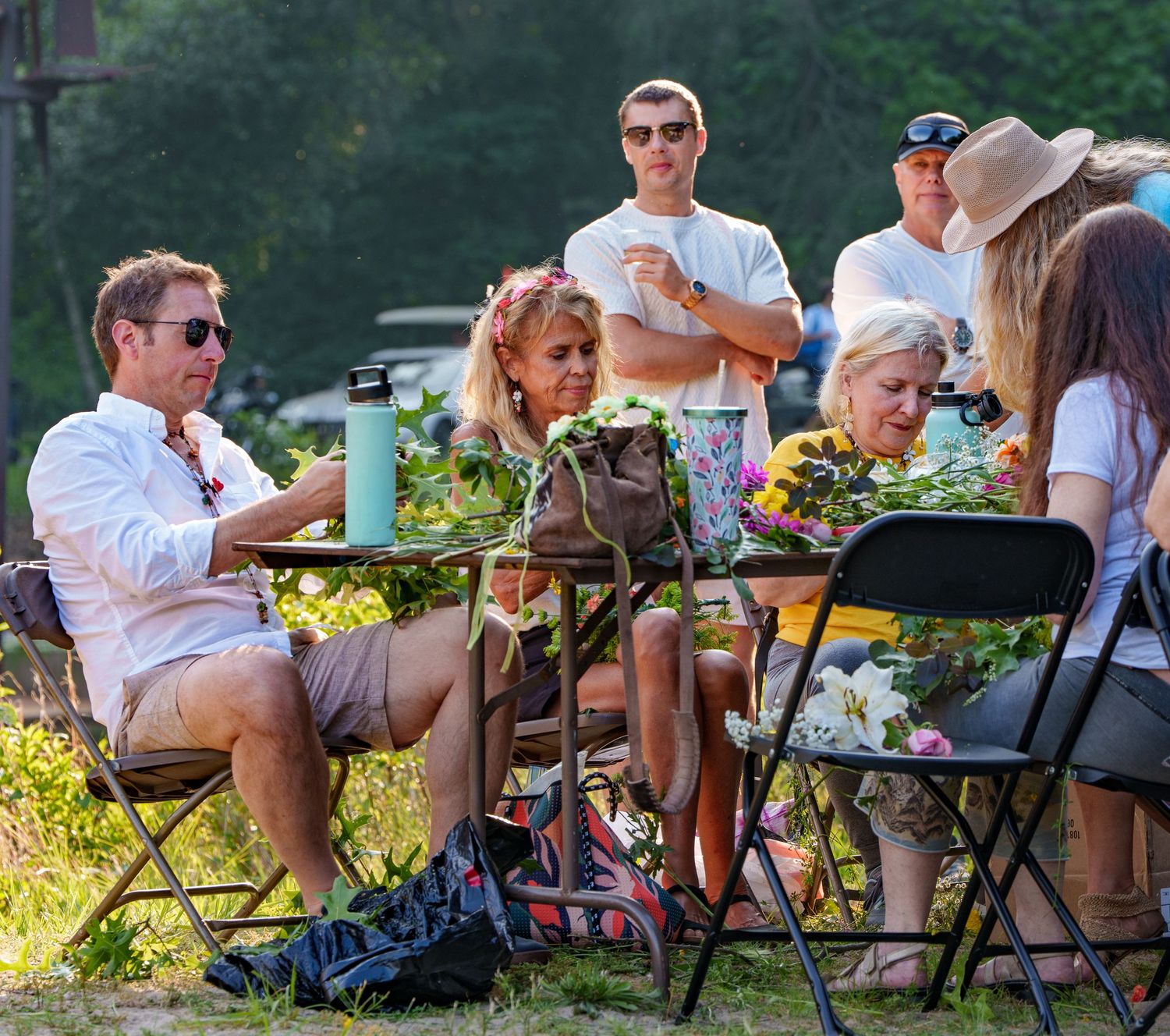 Group of people sitting and standing outdoors at a summer gathering with flowers on a table.