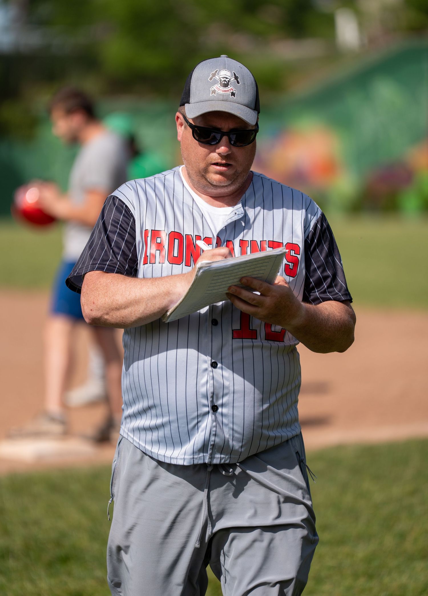 Baseball coach in pinstripe jersey reviews clipboard on sunny field during practice game.
