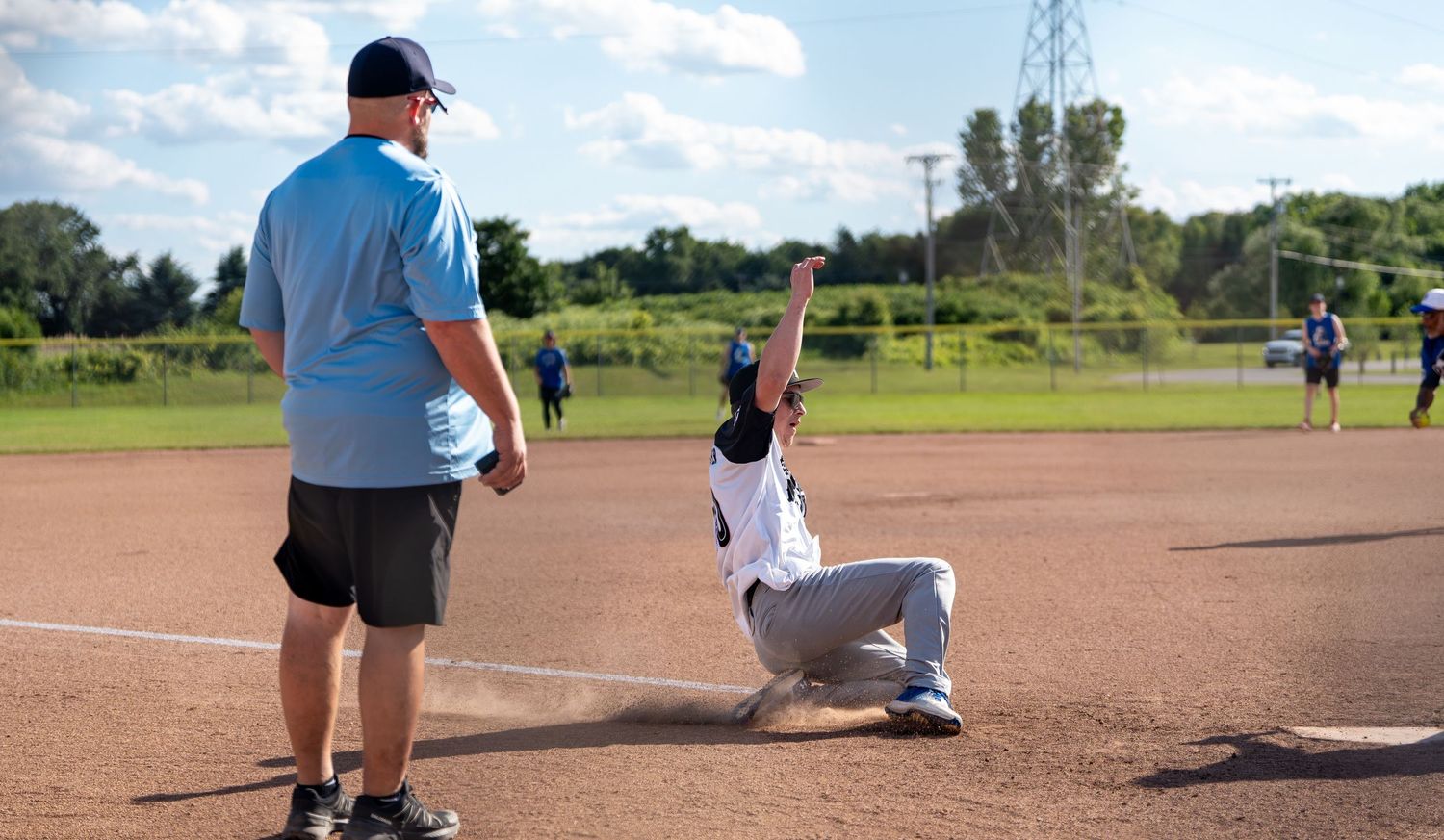 Baseball player sliding into base while coach watches during a sunny youth baseball game on a dirt field.