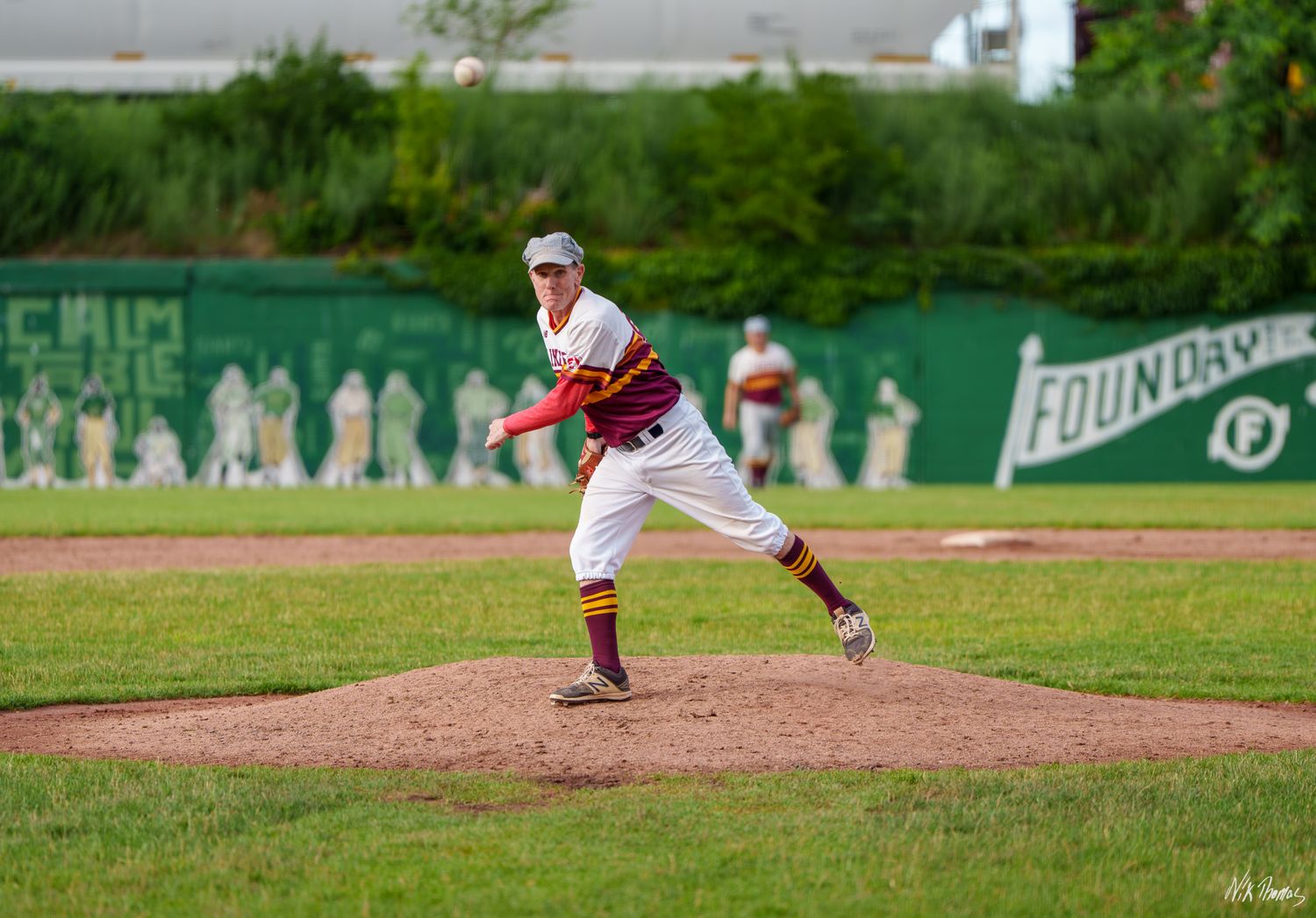 Baseball pitcher in white uniform winds up to throw on a green field mound during a sunny outdoor game.