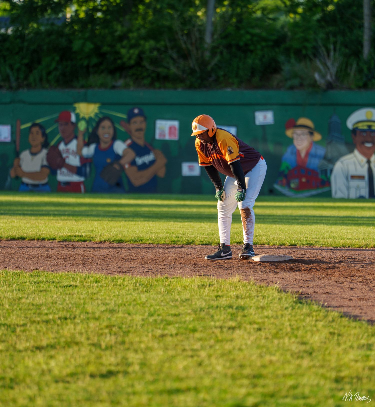 Baseball player in orange helmet crouches at base on green field with colorful mural in background.