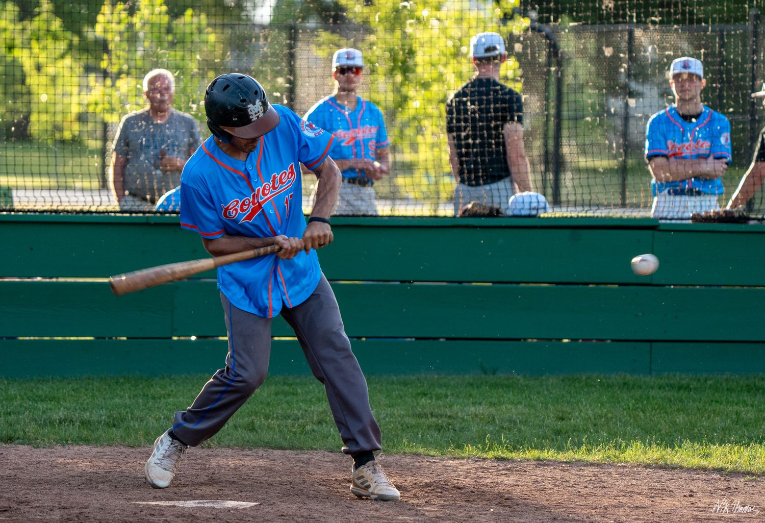 Baseball player in blue jersey swings bat at incoming pitch during practice game with teammates watching.