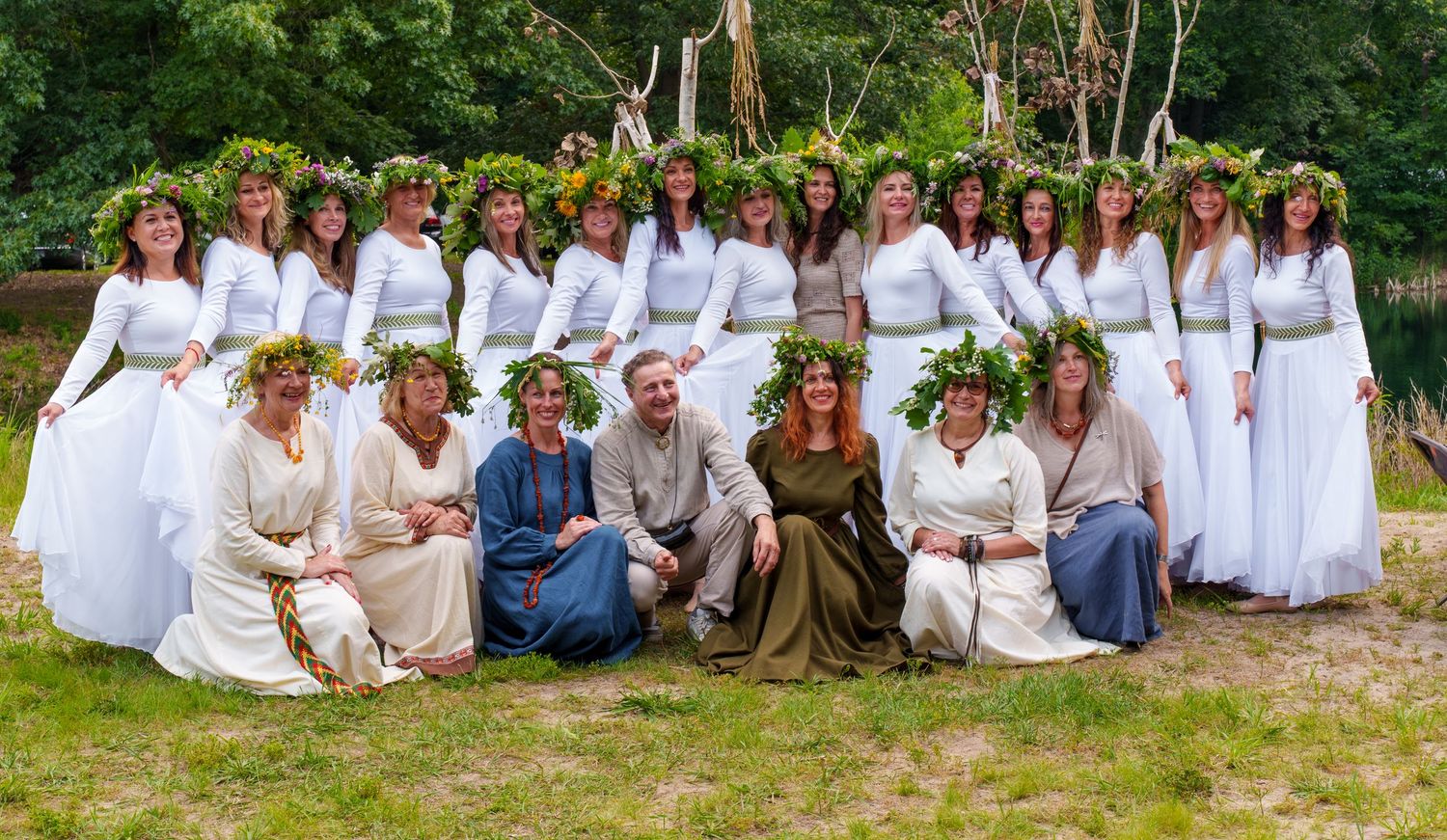 Large group photo of women in white dresses with floral wreaths at an outdoor folk festival.