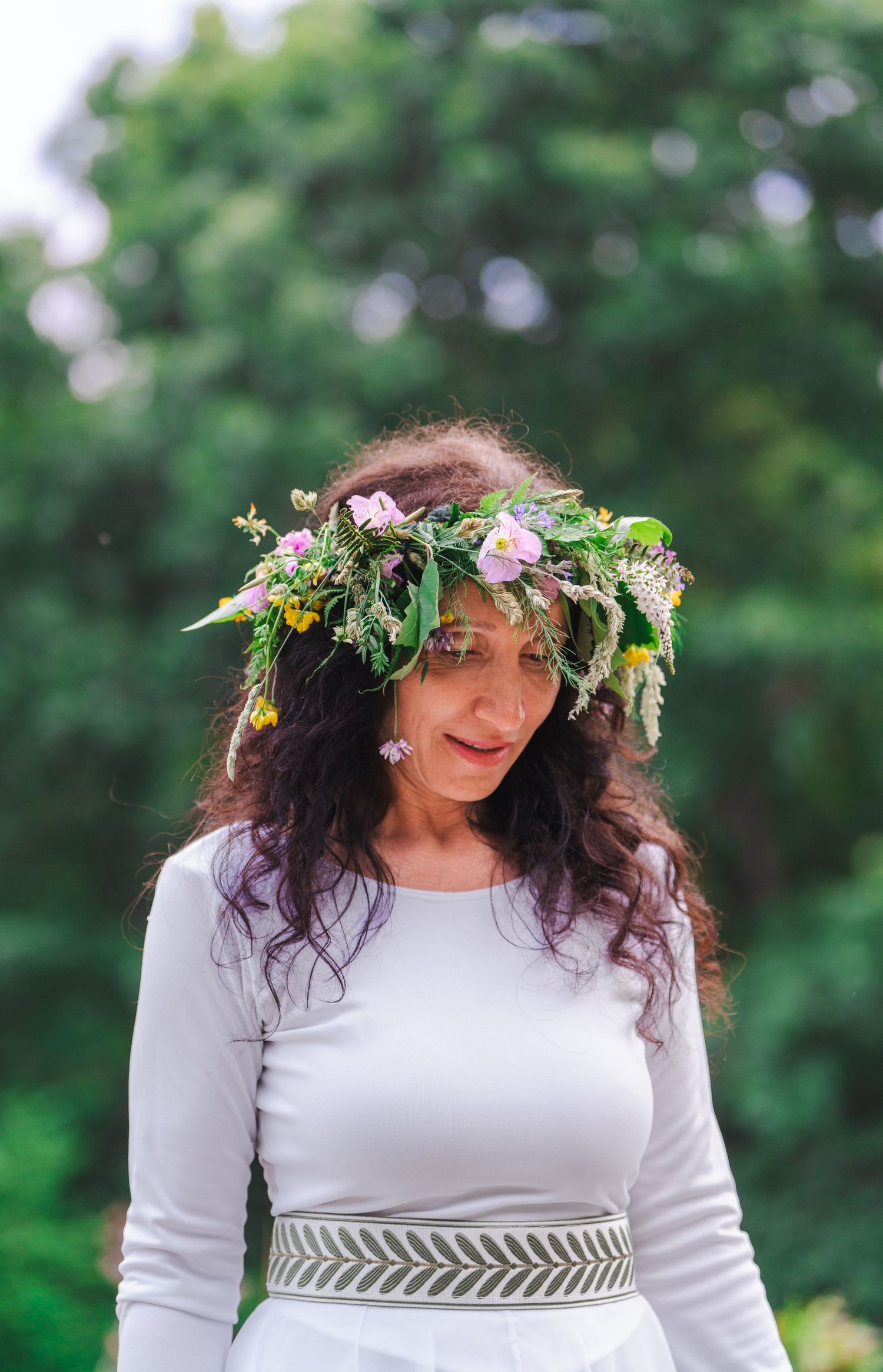 Woman wearing a floral crown of wildflowers in a white dress, standing outdoors with green trees in the background.