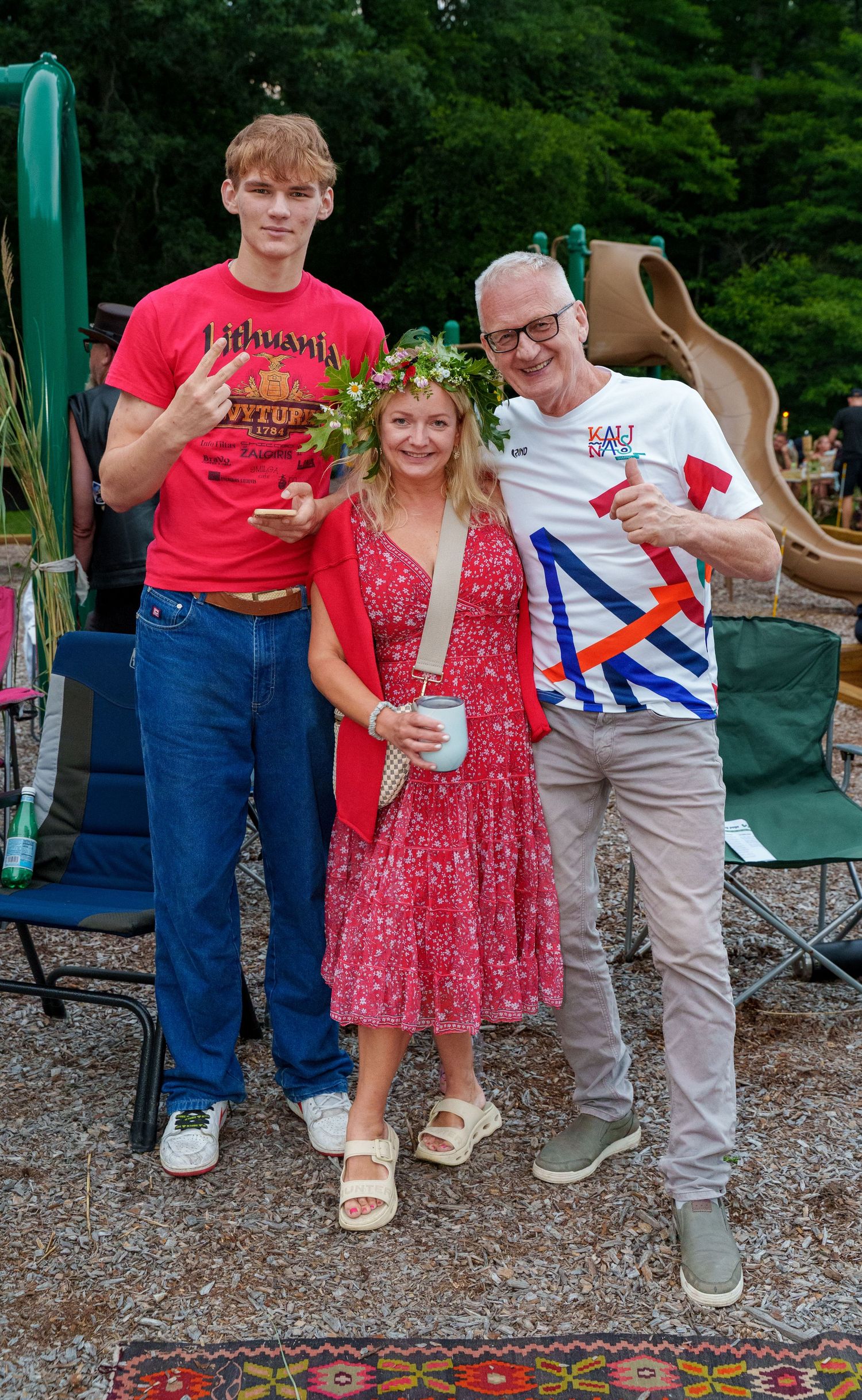 Three people pose outdoors at a summer festival, woman wearing flower crown in red dress flanked by two men.