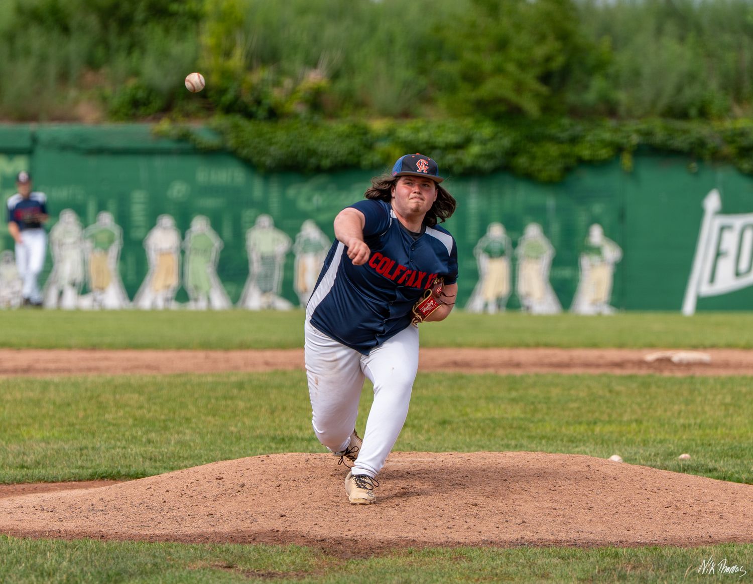 Baseball pitcher in navy uniform delivering a pitch from the mound during an outdoor game.