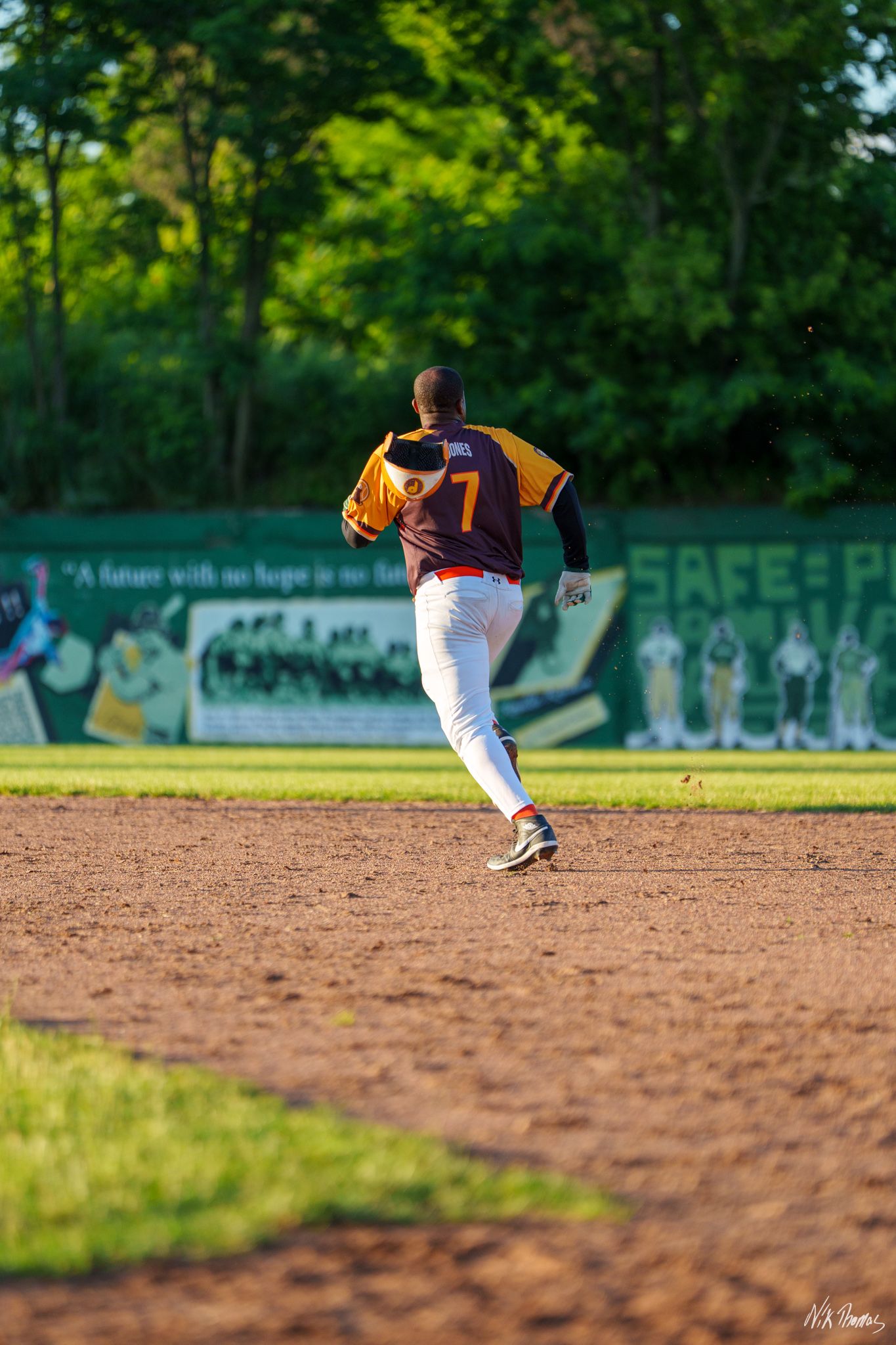 Baseball player wearing jersey number 7 runs the bases on a dirt infield with a green mural wall in the background.