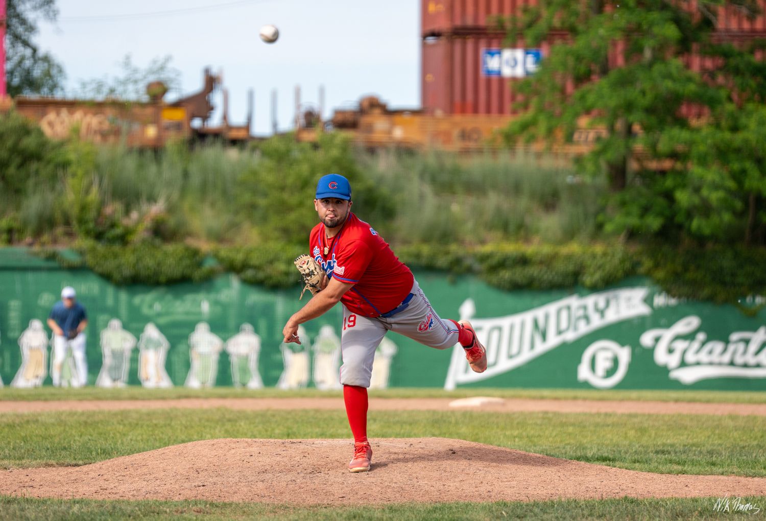 Baseball pitcher in red uniform mid-throw on mound during sunny game with green outfield wall visible.