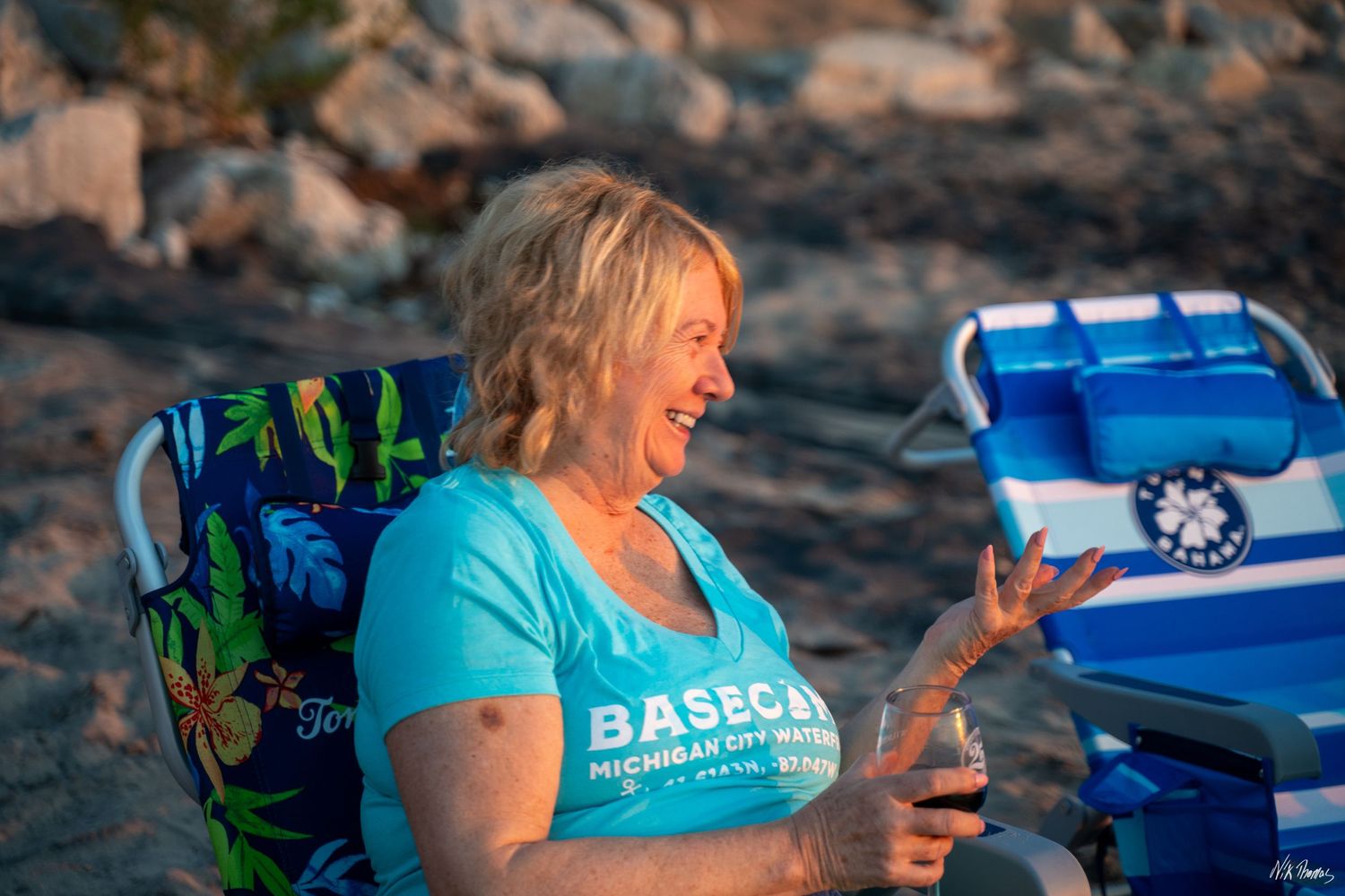 Smiling woman in teal BASECA Michigan shirt relaxing in beach chair on sandy shore at golden hour.