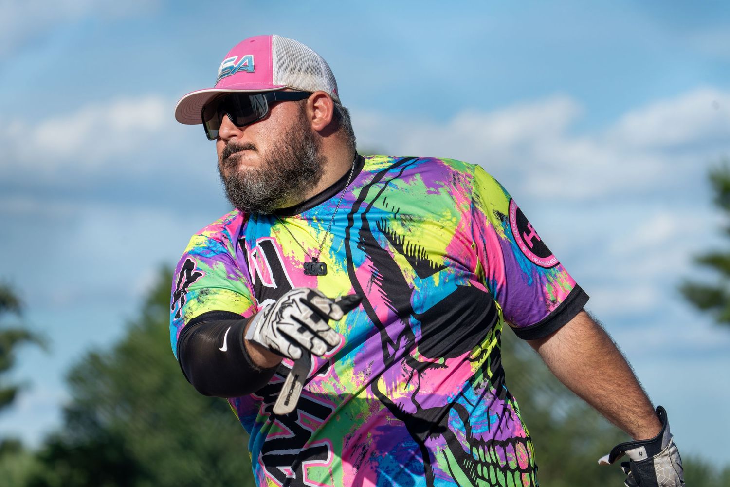 Man in colorful tie-dye jersey and cap playing softball outdoors on a sunny day.