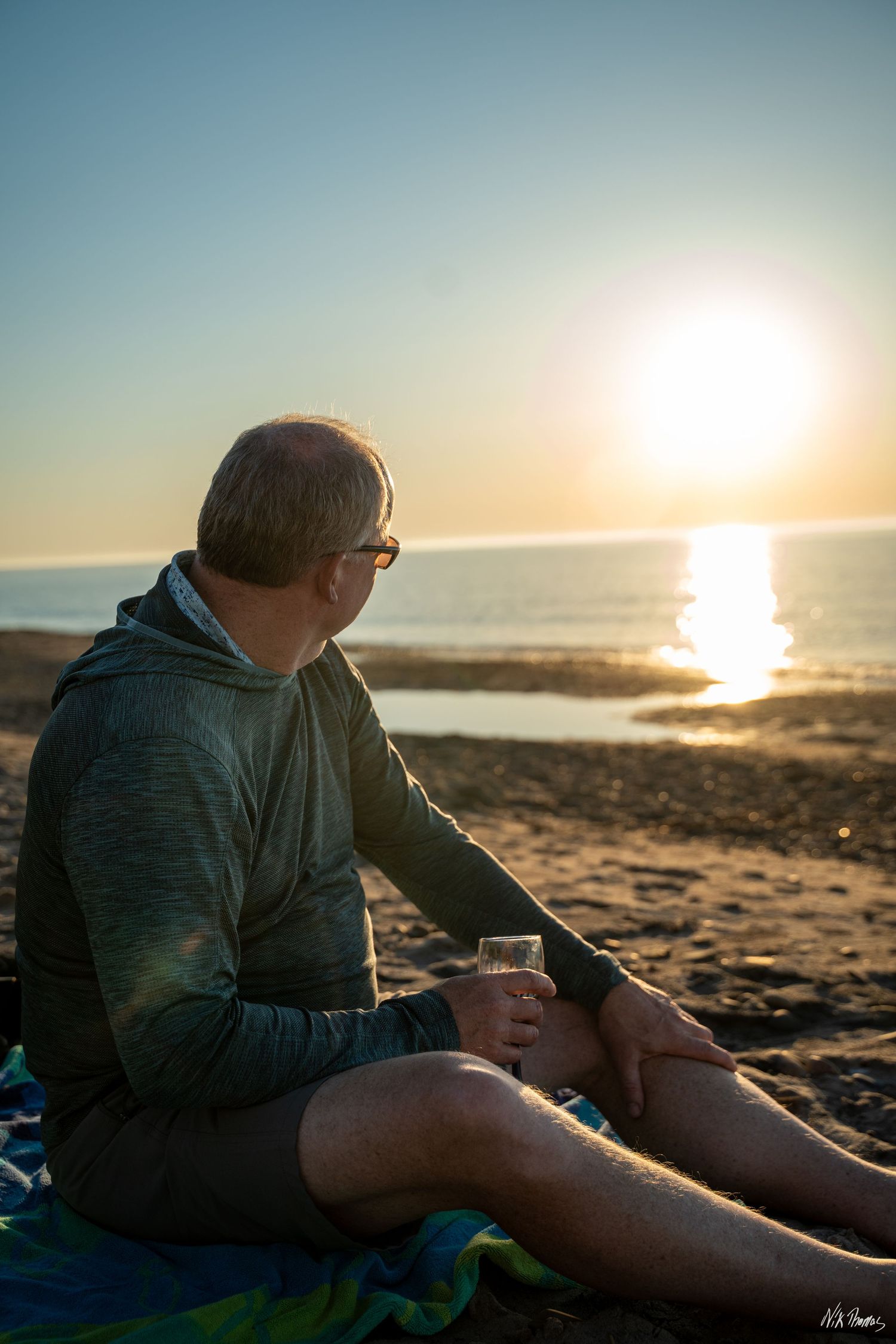 Man sitting on beach at sunset holding a drink, wearing glasses and green hoodie with shorts.