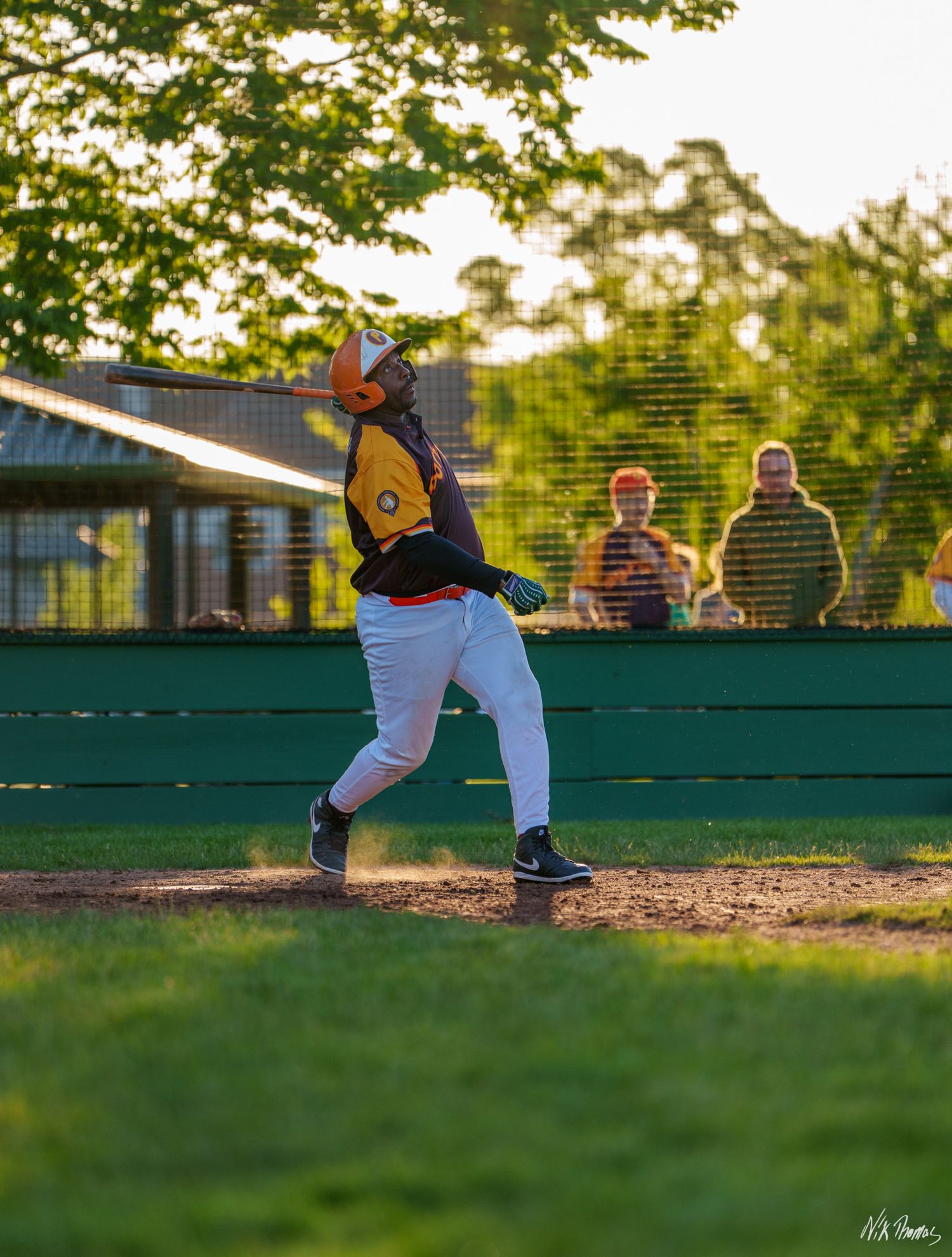 Youth baseball player in yellow jersey swings bat during evening game with spectators watching from behind green dugout fence.