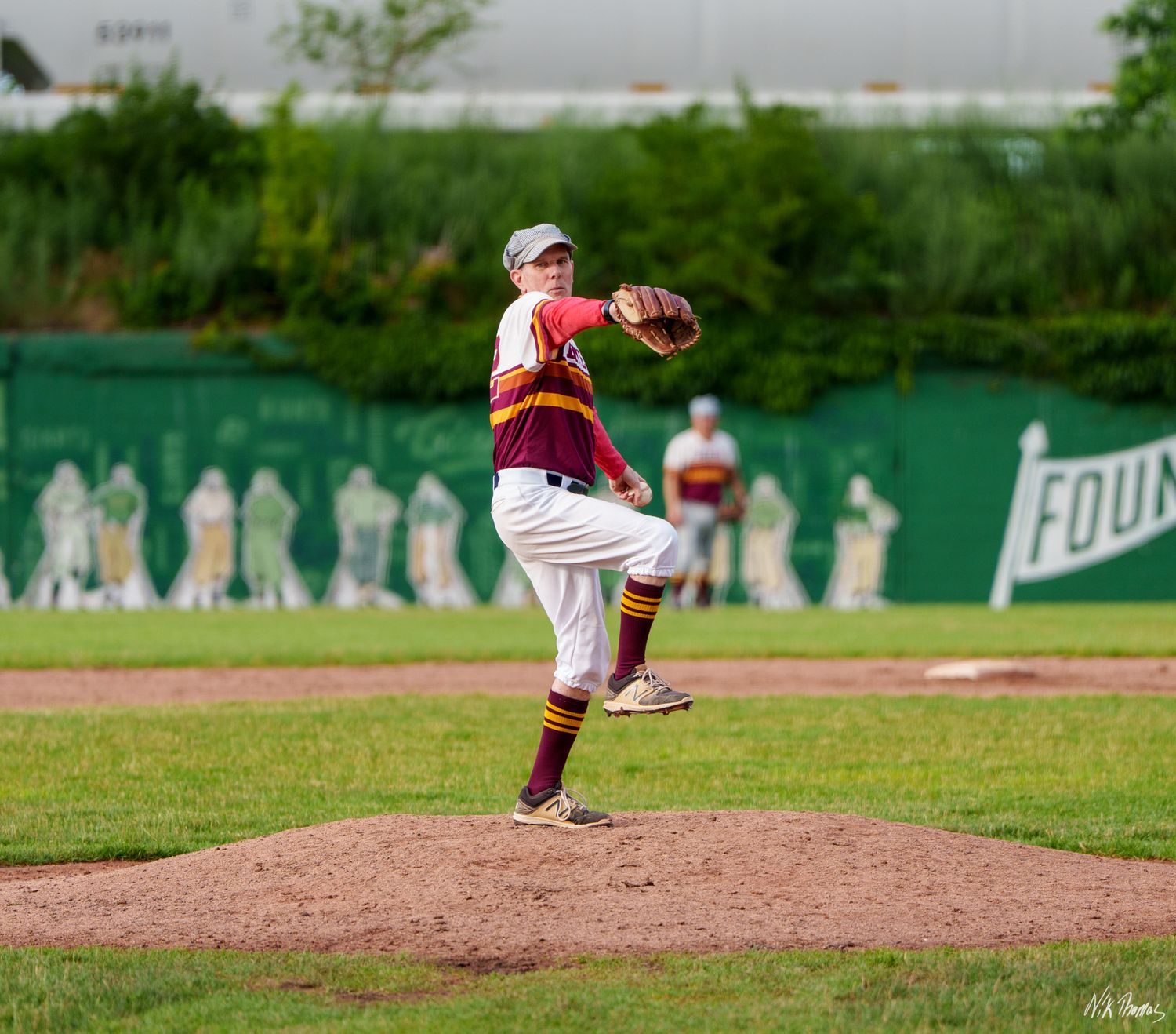 Baseball pitcher in maroon and gold uniform winds up to throw from the mound during an outdoor game.