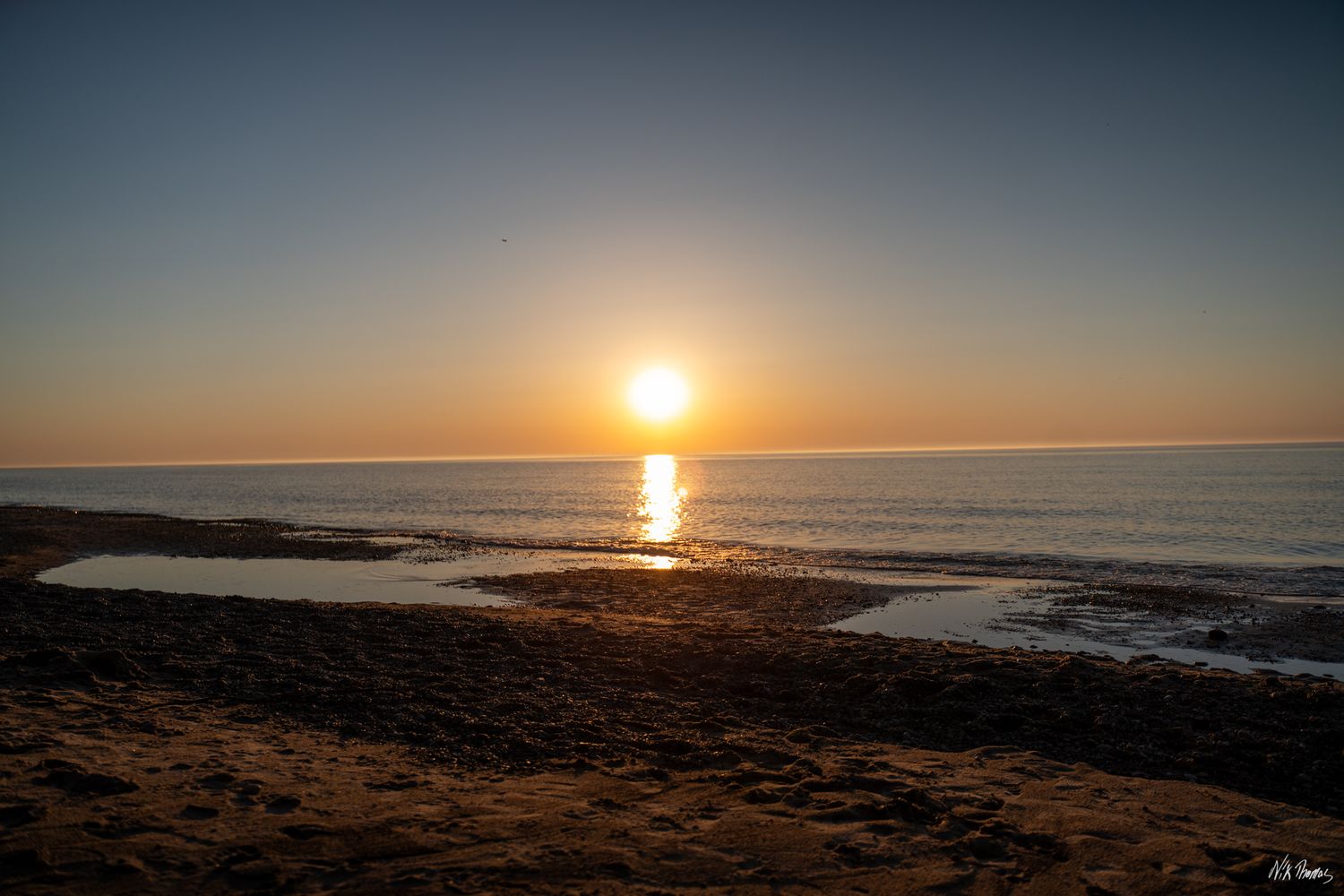 Golden sunset over calm ocean waters with sandy beach in foreground, reflecting warm orange hues on the horizon.