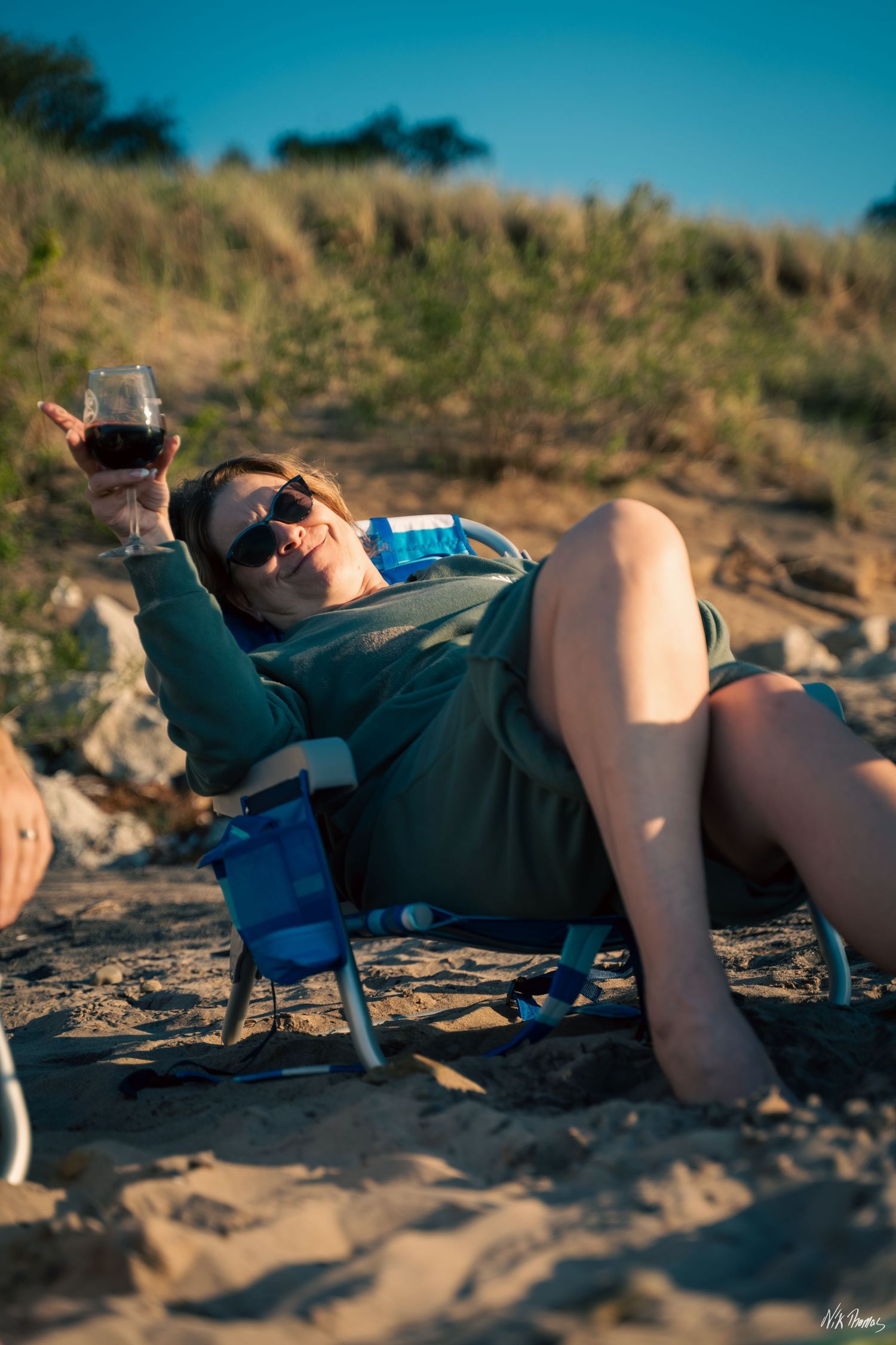 Woman relaxing in beach chair on sandy shore, holding red wine glass, wearing sunglasses and green outfit.