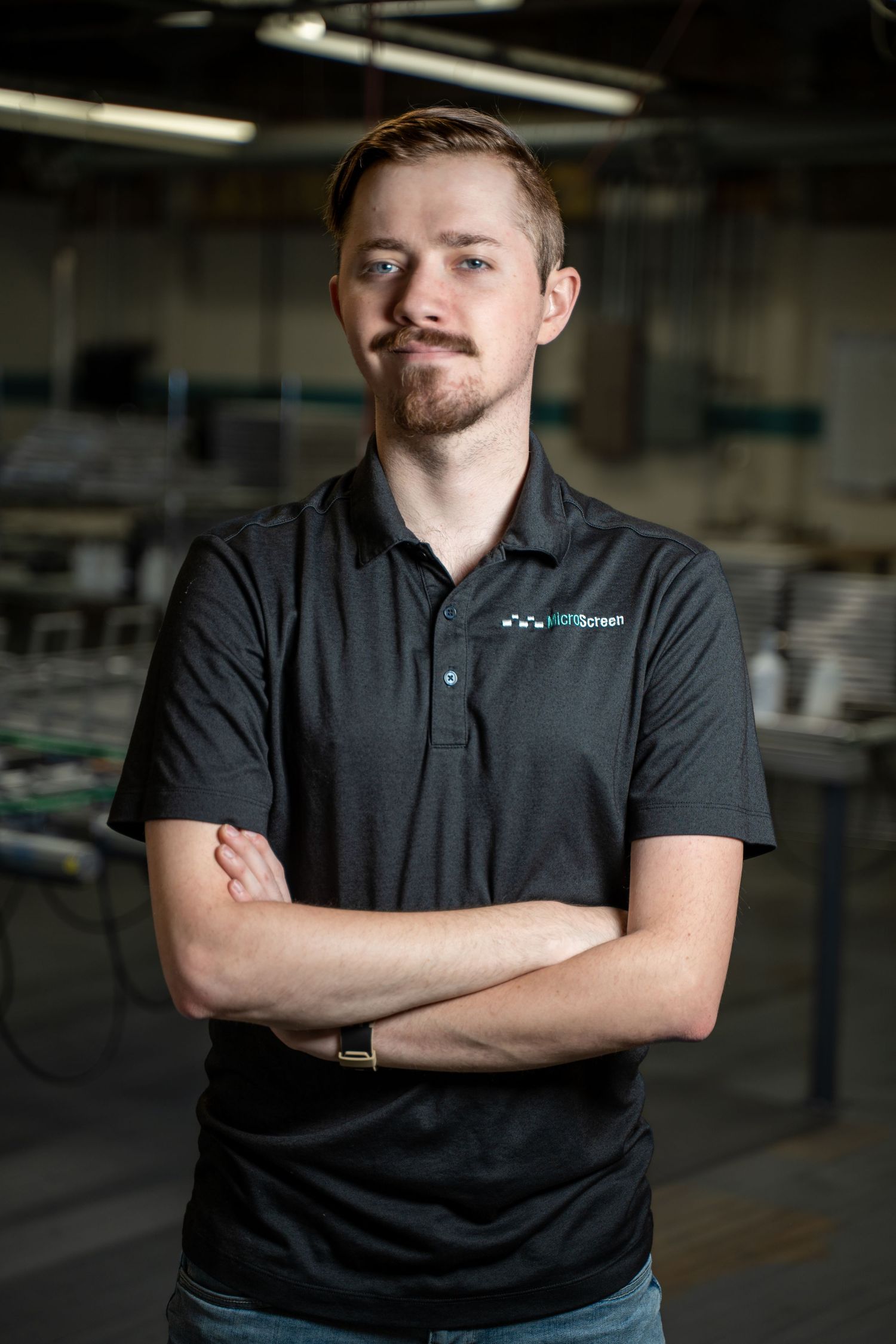 Young man with mustache stands arms crossed in dark polo shirt inside an industrial printing facility.