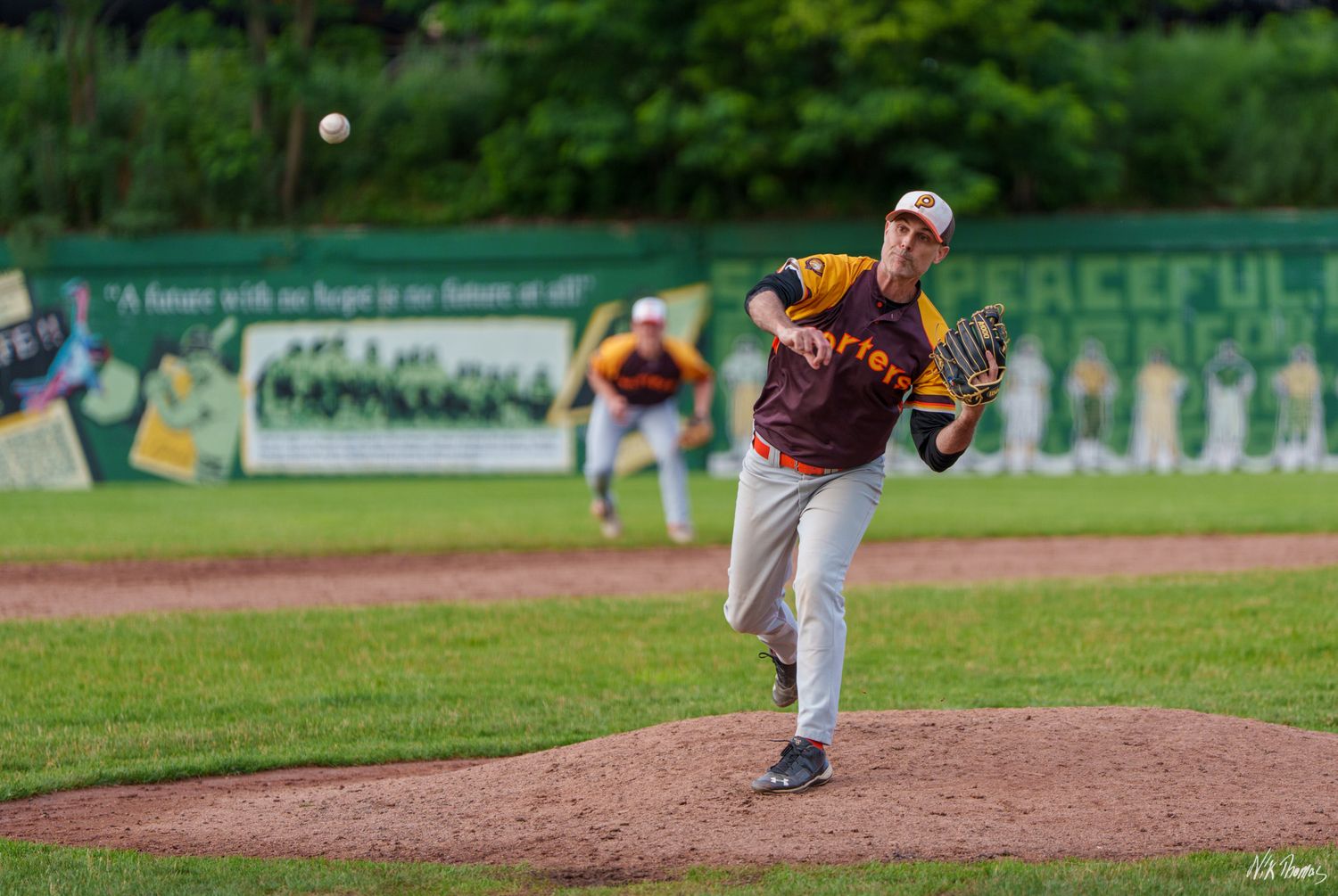 Baseball pitcher in maroon and gold uniform delivers a pitch during an outdoor game.