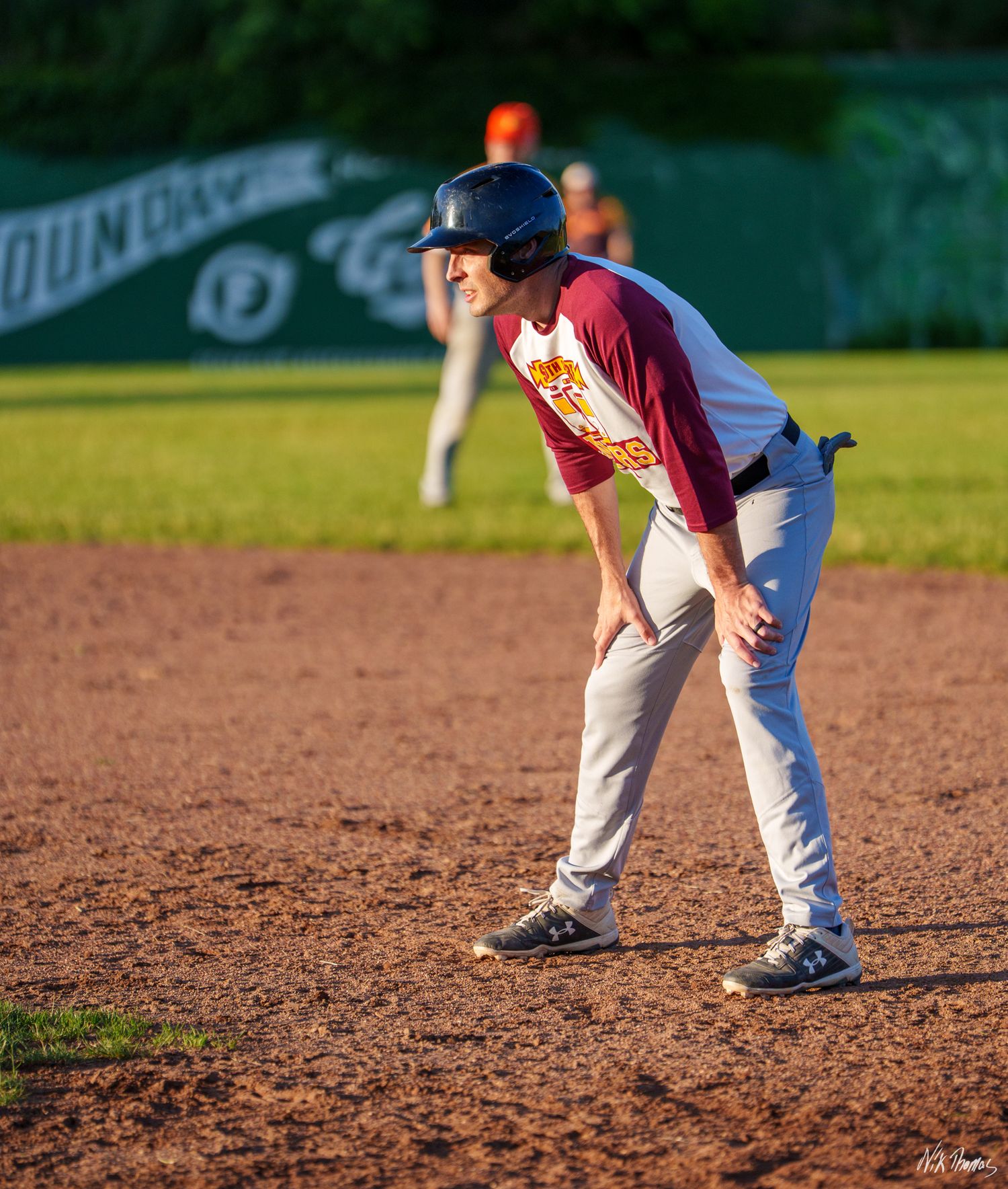 Baseball player in maroon uniform crouching at base during a sunny game on a dirt infield.