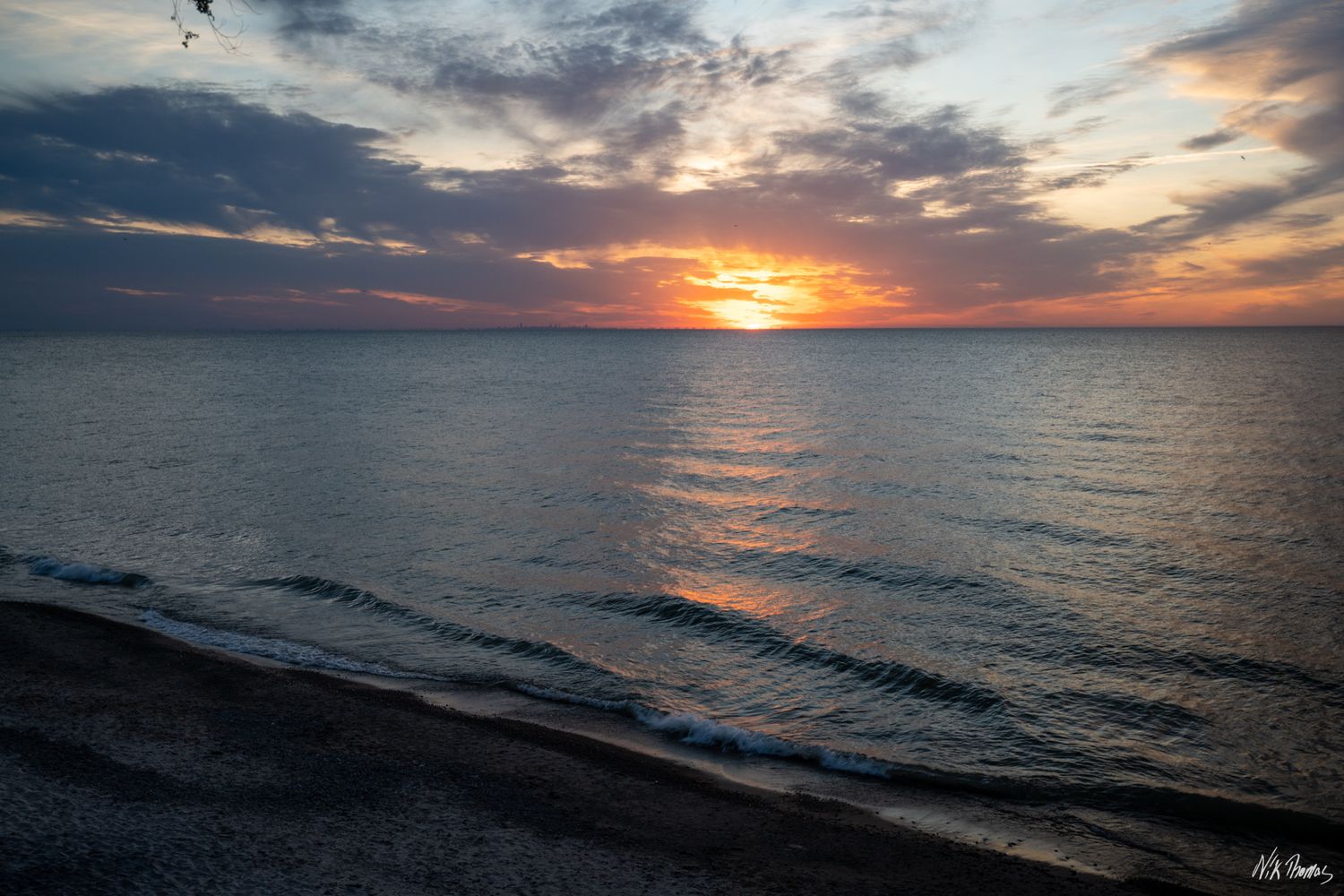 A stunning ocean sunset with vibrant orange and red hues reflecting on calm sea waters under dramatic clouds.