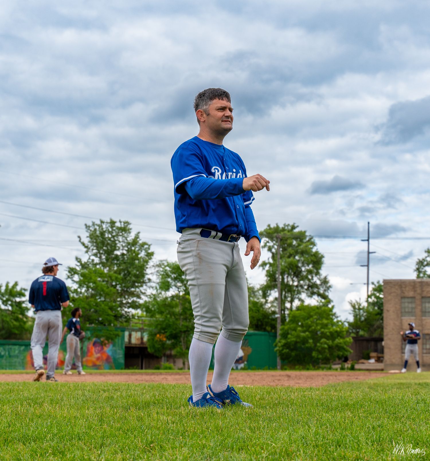 Baseball player in blue jersey and gray pants stands on green field during an outdoor baseball game.