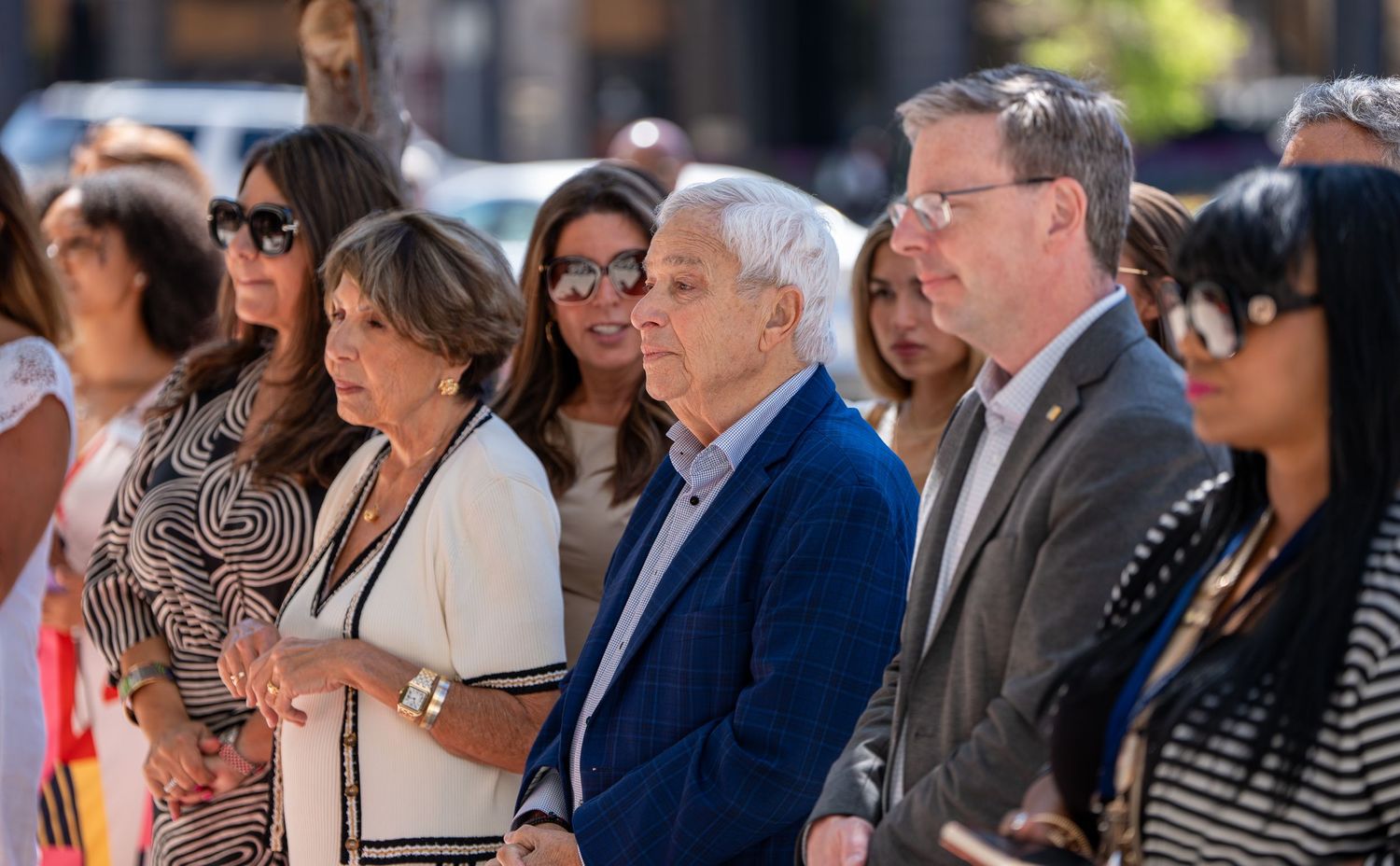 Group of formally dressed attendees standing together outdoors at a ribbon cutting ceremony.