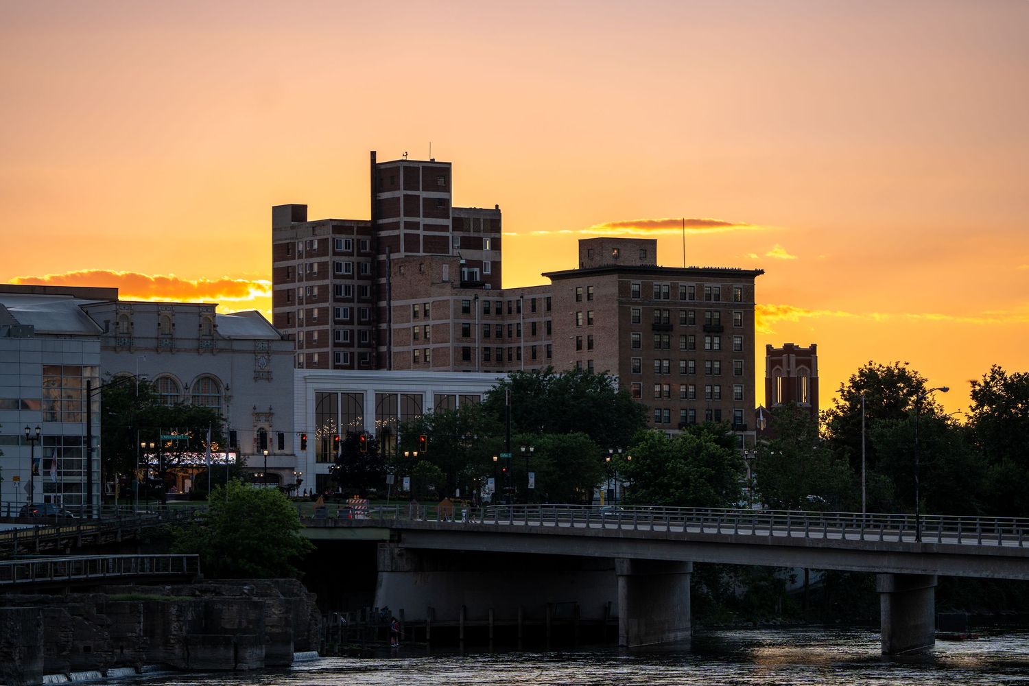 Downtown cityscape at golden hour sunset with bridge over river and historic brick buildings glowing orange.